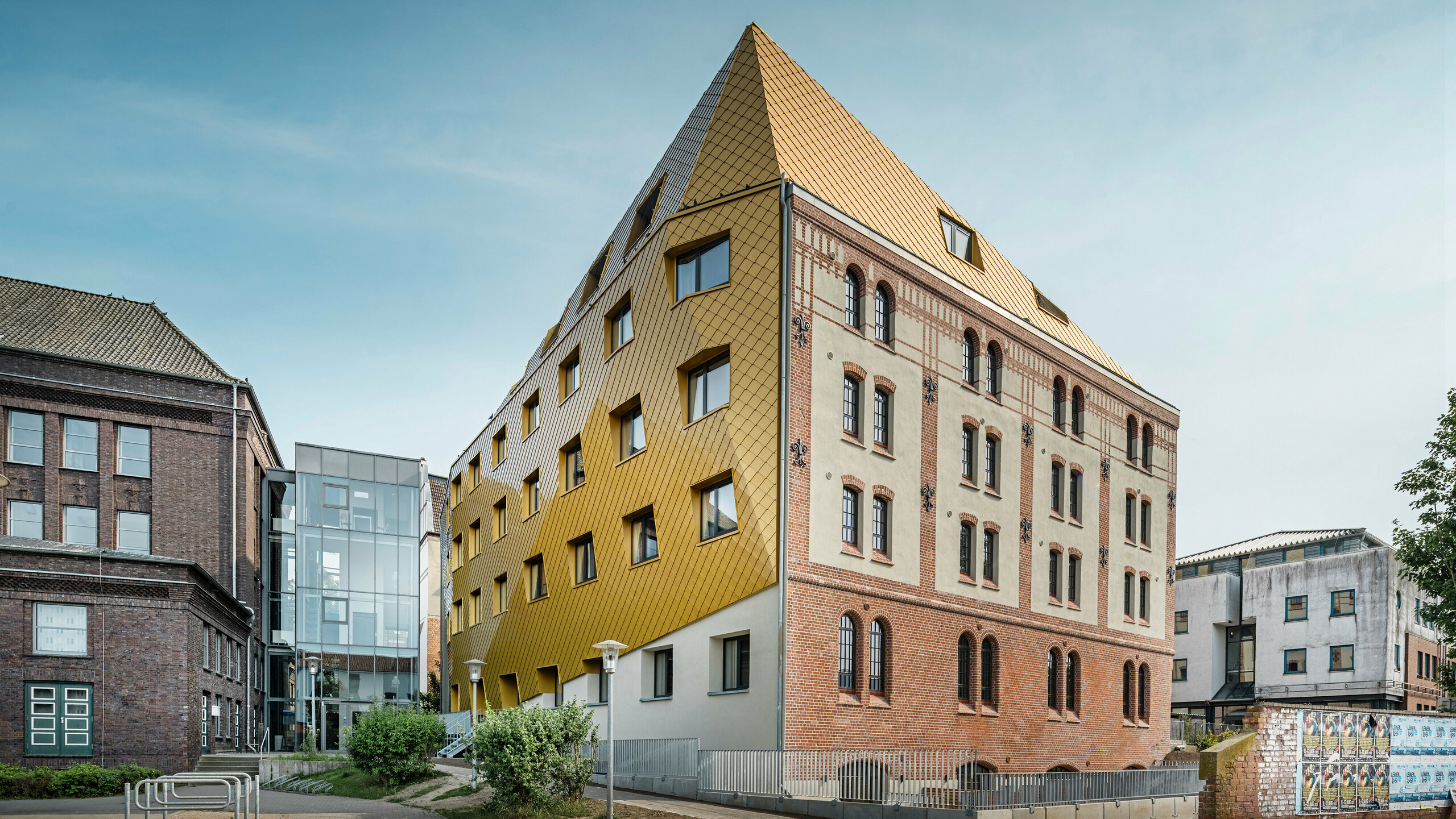 Bodenaufnahme auf "The Station" in Kiel. Das Studentenwohnheim ist schräg von der Seite zu sehen. Auf dem Bild wird der eklatante Kontrast zwischen der Aluminiumfassade aus PREFA Dach- und Wandrauten 29 × 29 in Mayagold auf der linken Seite und der denkmalgeschützten Putz- und Ziegelfassade auf der rechten Seite deutlich.