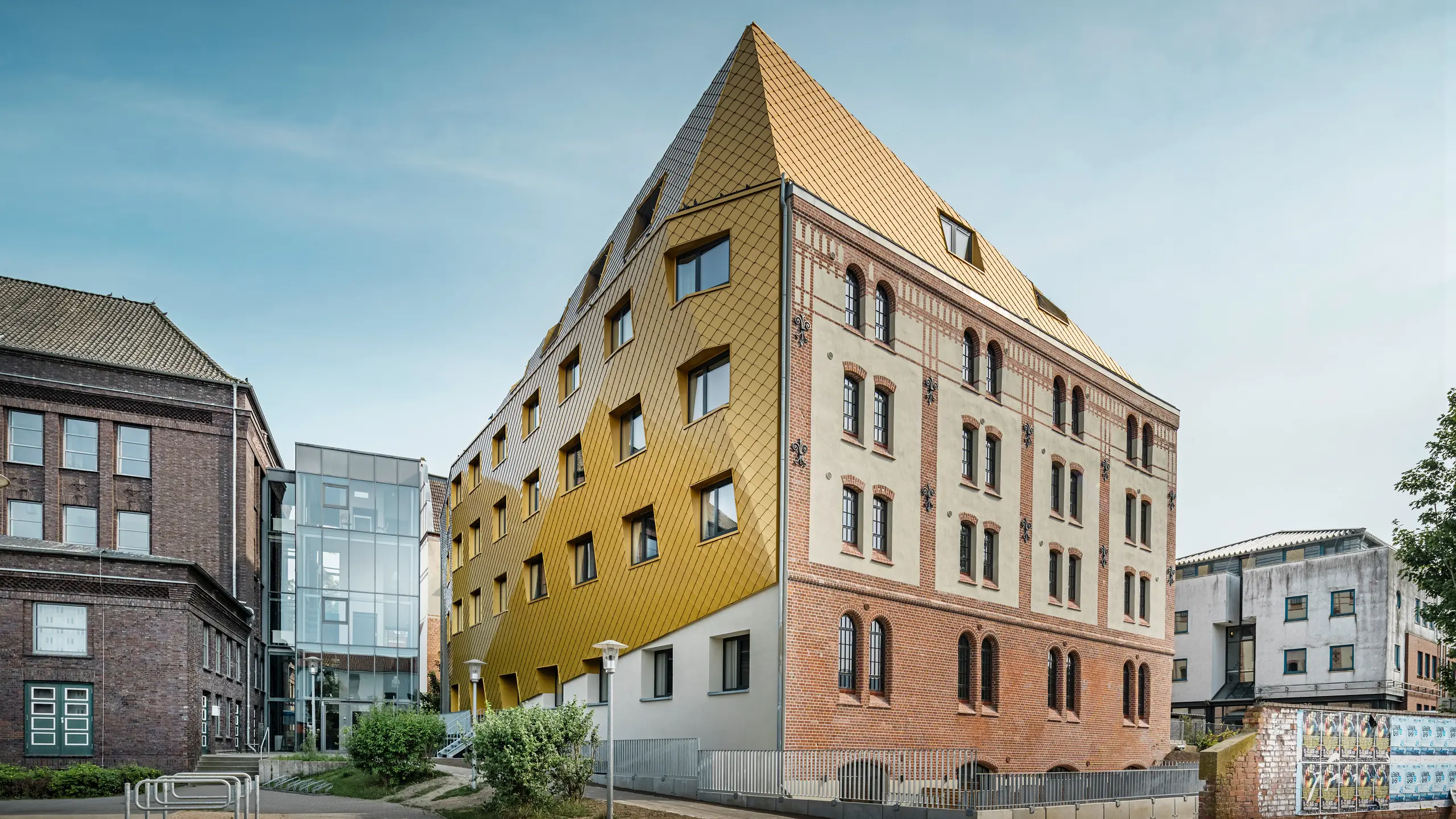 Bodenaufnahme auf "The Station" in Kiel. Das Studentenwohnheim ist schräg von der Seite zu sehen. Auf dem Bild wird der eklatante Kontrast zwischen der Aluminiumfassade aus PREFA Dach- und Wandrauten 29 × 29 in Mayagold auf der linken Seite und der denkmalgeschützten Putz- und Ziegelfassade auf der rechten Seite deutlich.