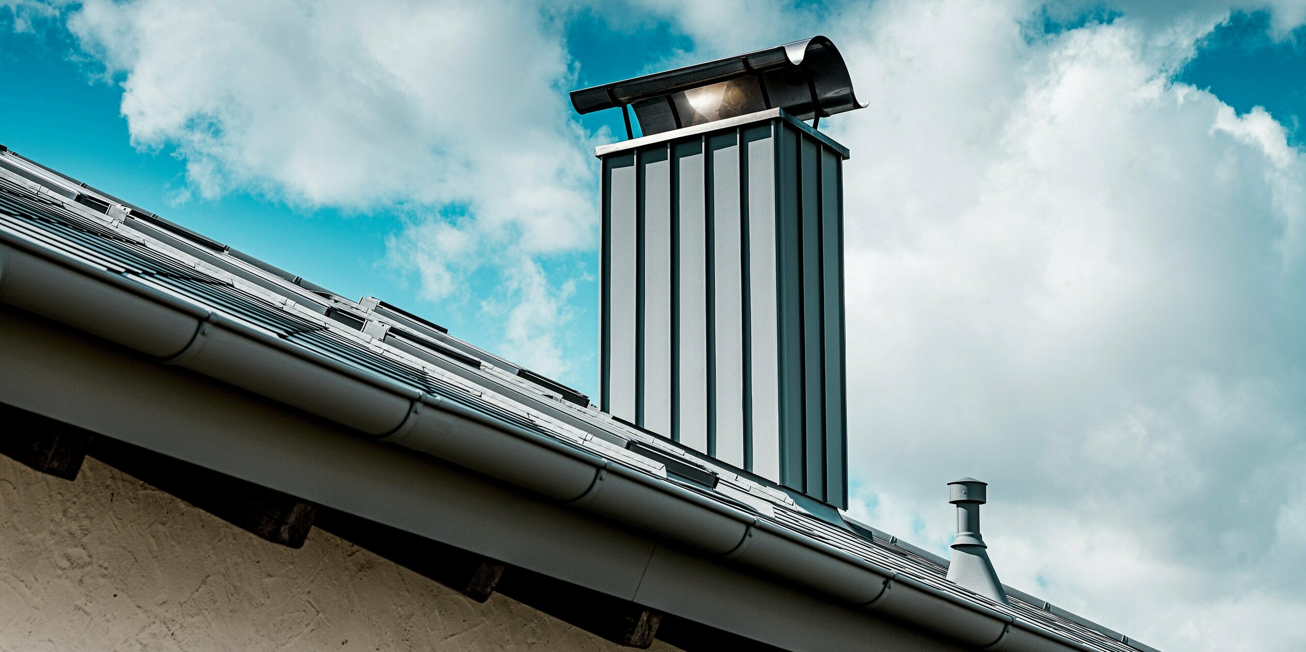 Das Bild zeigt eine Nahaufnahme des modern verkleideten Kamins eines Einfamilienhauses, der mit hochwertigem PREFA Aluminium in P.10 Hellgrau gestaltet ist. Die klaren vertikalen Linien der Kaminverkleidung harmonieren mit der robusten PREFA Dachfläche aus Dachrauten 44 × 44. Im Vordergrund ist die widerstandsfähige Aluminium-Dachrinne sichtbar, die sich nahtlos in das Dachsystem einfügt. Vor dem Hintergrund eines wolkigen Himmels hebt sich die langlebige, witterungsbeständige PREFA Lösung besonders markant ab.