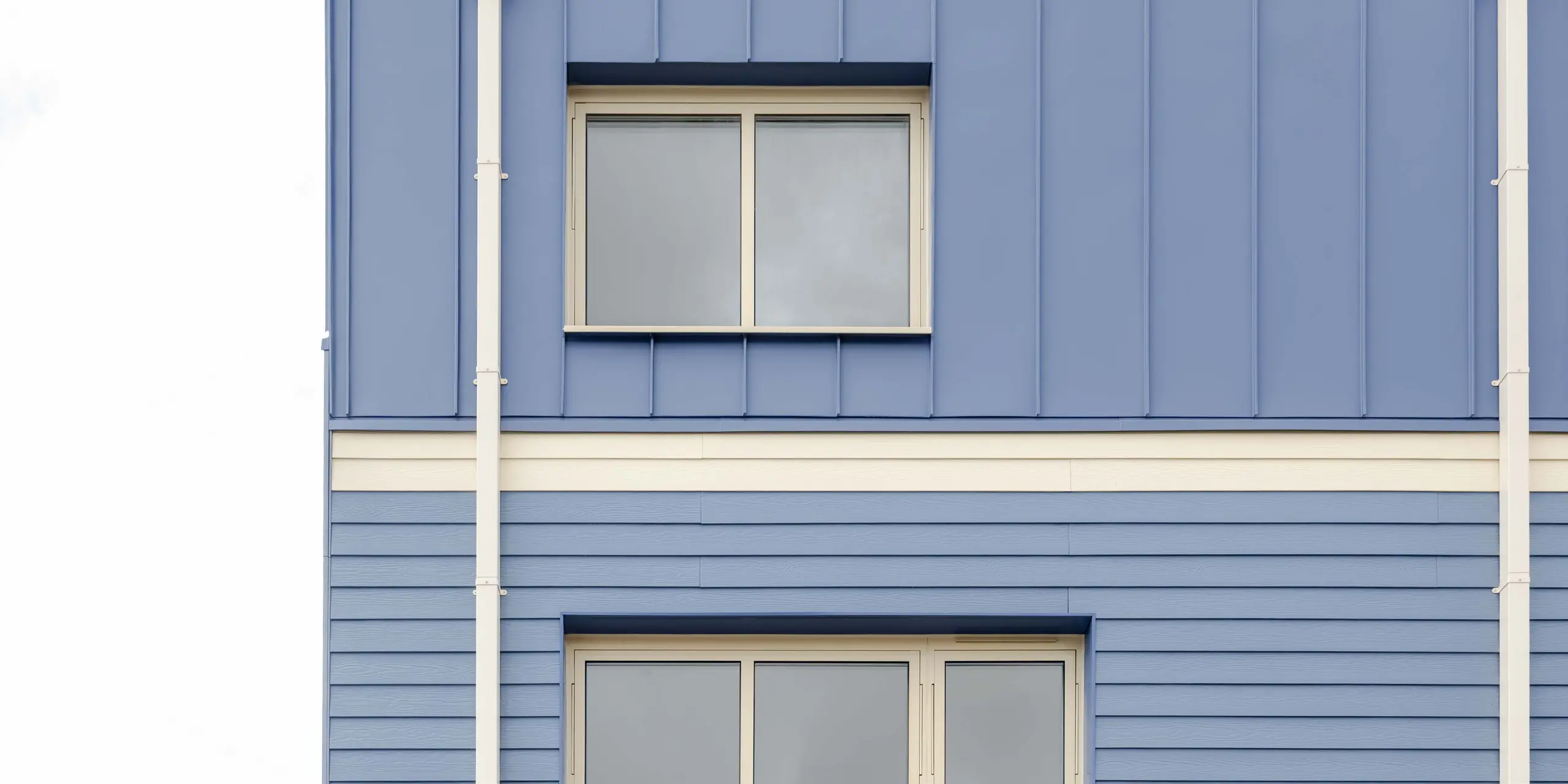 Die Aufnahme zeigt die Rückseite eines Goldsmith‑Mews‑Reihenhauses mit der markanten PREFA FALZONAL Verkleidung in Taubenblau. Die klar strukturierten, vertikalen Aluminiumfalze im oberen Bereich treffen auf die horizontalen Paneele des Mittelgeschosses und erzeugen ein stark gegliedertes Fassadenbild. Die hellen unteren Bereiche mit großen Terrassentüren öffnen die Architektur zum Garten. Das hochwertige PREFA Aluminium bietet hohe Farb- und Formstabilität, dauerhaften Witterungsschutz und ermöglicht eine moderne, präzise gestaltete Gebäudehülle mit klarer Wiedererkennung.