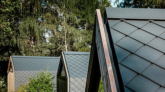 Mehrere minimalistische A‑Frame‑Tiny‑Houses des Secret Koi Garden reihen sich harmonisch am Waldrand des Natterer Sees aneinander. Die Dachflächen sind mit kleinteiligen PREFA Rauten aus Aluminium in P.10 Anthrazit gedeckt und erzeugen eine markante, fischhautähnliche Struktur. Die matte Oberfläche wirkt ruhig und elegant, während das langlebige, witterungsbeständige PREFA Aluminium optimalen Schutz in alpiner Umgebung bietet und Design und Natur perfekt verbindet.