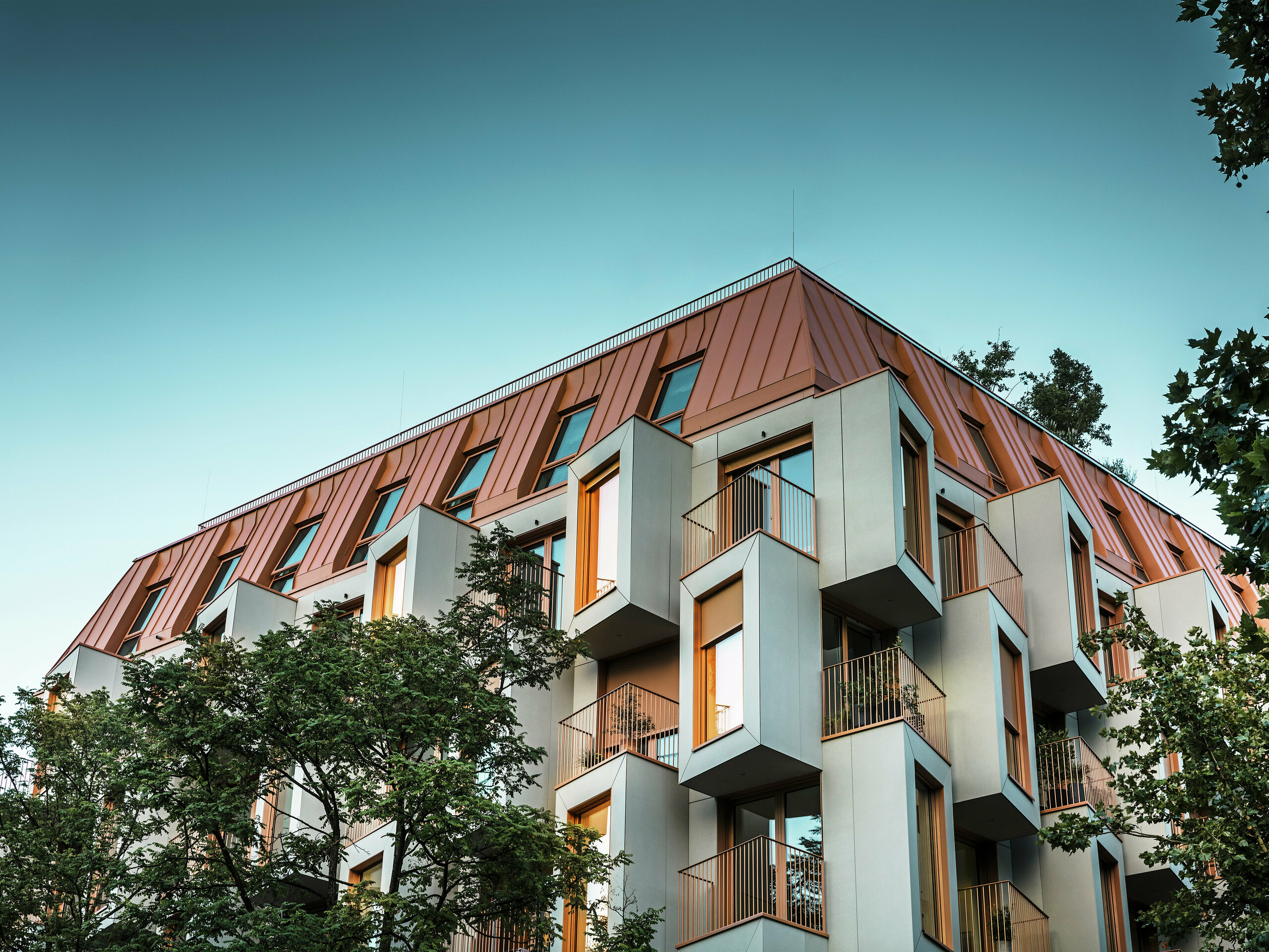 Architektonisch herausragende Fassade des Wohnprojektes Van B in München mit markanten Erkern und PREFALZ Aluminiumdach in der Sonderfarbe IGP Orange HDP. In der Dämmerung zeigt der Blick von unten die Lichter in den Fenstern. Die moderne Dachlösung verbindet höchste Witterungsbeständigkeit mit eleganter Optik und setzt farbliche Akzente im Stadtraum. Das Zusammenspiel von Glasfaserbeton, kupferorangen Fensterrahmen und Aluminiumdach schafft eine dynamische, langlebige und nachhaltige Gebäudehülle - ideal für zukunftsorientiertes Bauen.