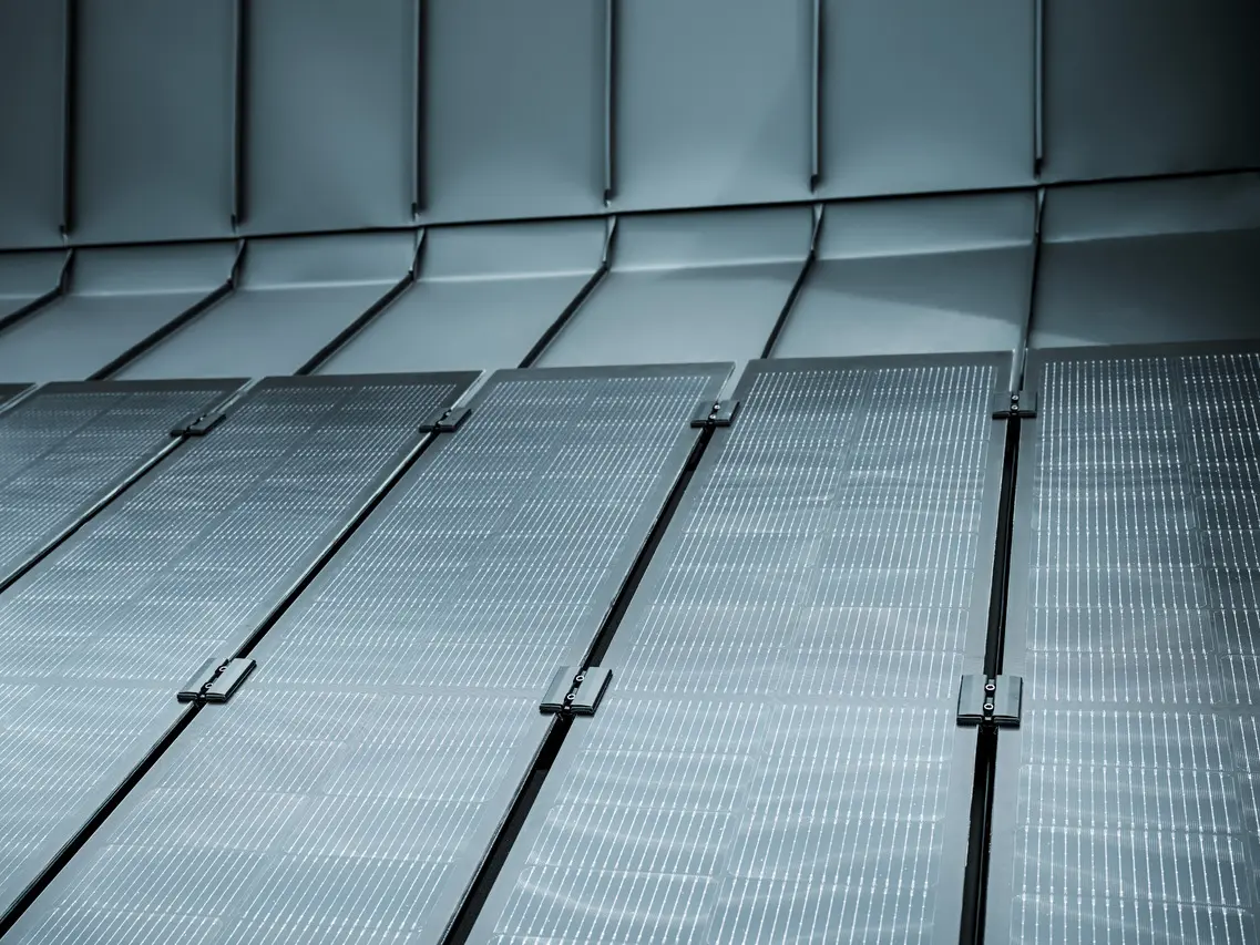 Detailaufnahme einer modernen PREFALZ Dachfläche mit integrierten Solarmodulen. Die rahmenlosen Glas/Glas-PV-Module mit innovativer TOPCon-Zellentechnologie fügen sich nahtlos in das elegante Aluminiumdach ein und sorgen für eine ästhetische, homogene Optik. Die dunkle Oberfläche reflektiert das Licht dezent und unterstreicht das hochwertige Design. Dank flacher Neigung profitieren die Module von einem verbesserten Selbstreinigungseffekt, was langfristig maximale Effizienz und geringen Wartungsaufwand garantiert – eine perfekte Kombination aus Nachhaltigkeit, Leistung und Design.
