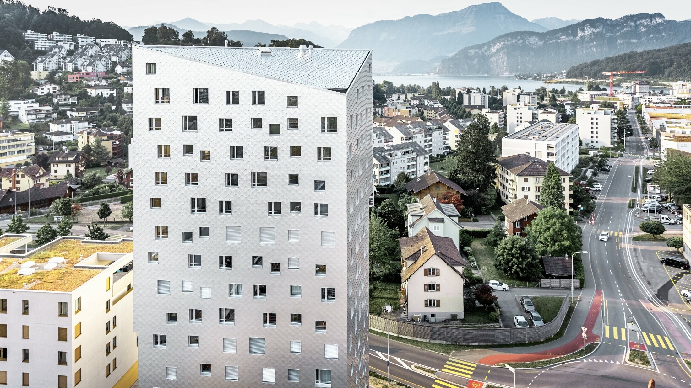 Das Bild zeigt das Solitaire-Hochhaus in Horw, einem Vorort von Luzern, das von der Schweizer Architektin Tilla Theus entworfen wurde. Das Hochhaus hat eine Fassade aus eloxierten Aluminiumrauten, die die Umgebung und die Wolken reflektieren. Die Fenster sind unregelmäßig angeordnet und als 'tanzende Fenster' bekannt. Im Hintergrund sind weitere Gebäude, Straßen und die malerische Berglandschaft der Schweiz zu sehen. Das Hochhaus steht inmitten einer städtischen Umgebung mit einer Mischung aus Wohnhäusern und städtischer Infrastruktur.