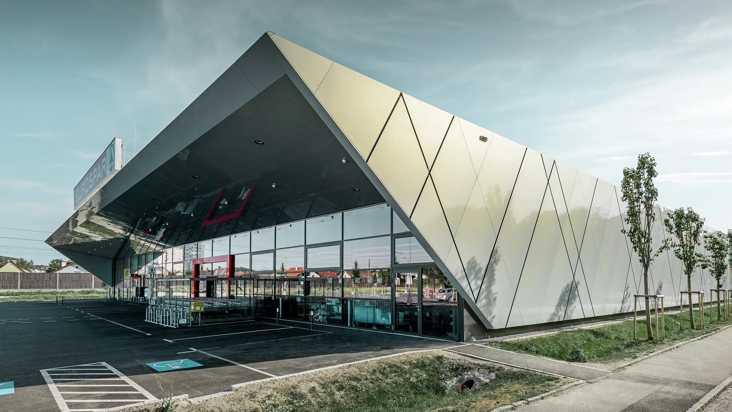 Der Eingangsbereich des EUROSPAR Supermarkts kombiniert großzügige Glasflächen mit einer markanten PREFA Aluminiumfassade. Die glänzende Oberfläche der Sonderfarbe Silver Gold unterstreicht die hochwertige, zukunftsorientierte Architektur der Supermarktfiliale. Die Aluminium Verbundplatten schillern in der Sonne je nach Blickwinkel in unterschiedlichen Farben.