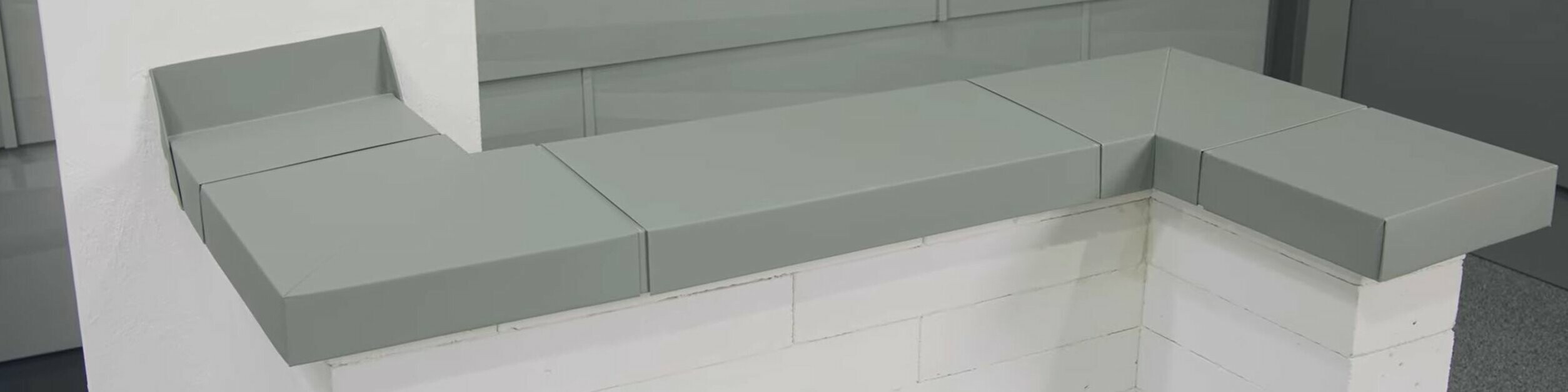 Graue PREFA Mauerabdeckung aus Aluminium auf weißem Mauerwerk mit moderner Fassadenverkleidung im Hintergrund