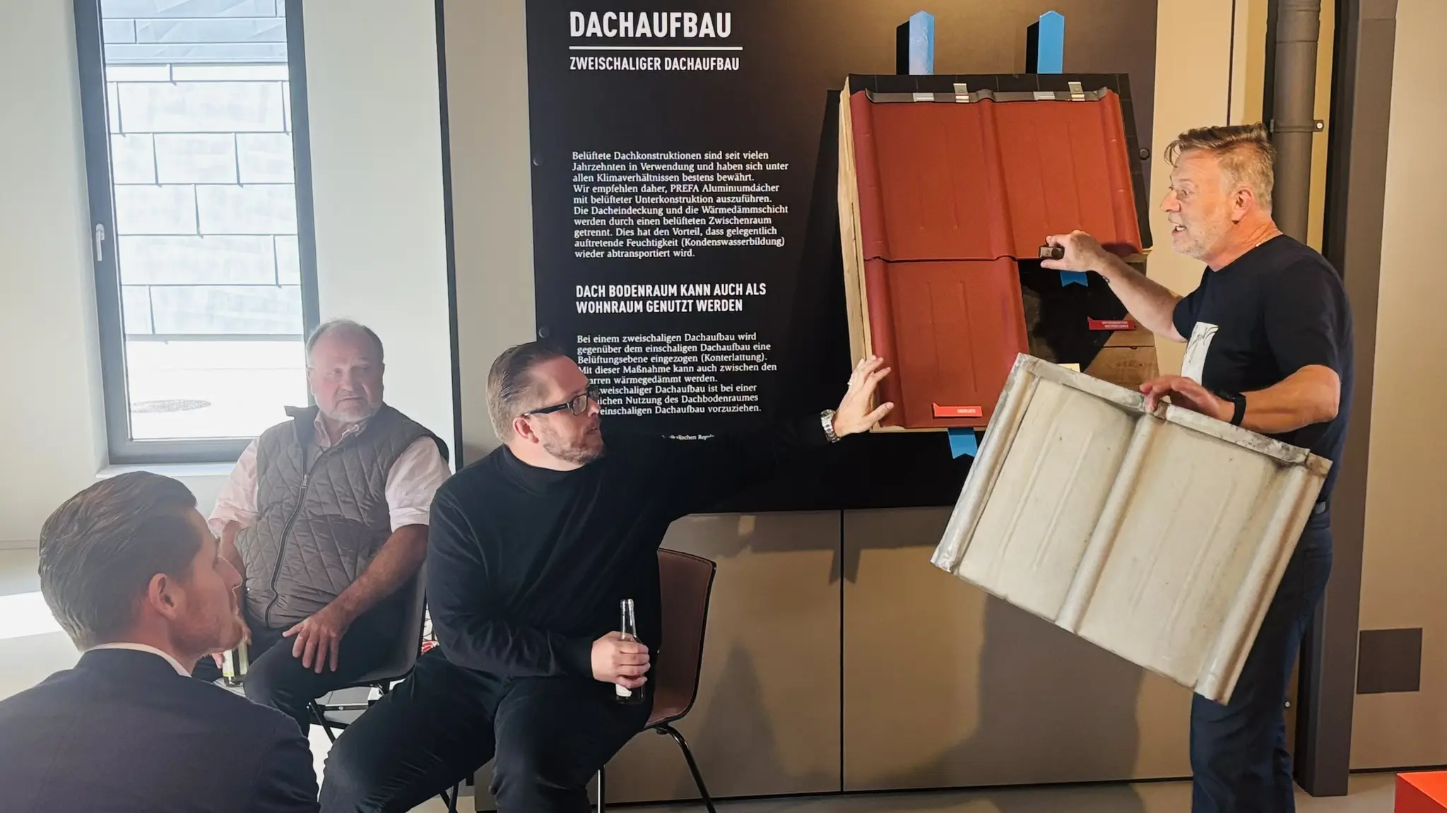 Im PREFA Showroom St. Veit an der Glan demonstriert Ralf Stadlober, PREFA Objektberater, anschaulich den zweischaligen Dachaufbau mit PREFA Aluminium-Dachsystemen. Eine Muster-Dachplatte sowie ein Querschnitt werden vor einer kleinen Gruppe erklärt, während Teilnehmende aufmerksam zuhören. Im Hintergrund informiert eine Tafel über den „Dachaufbau“ und dessen technische Eigenschaften. Die Szene zeigt praxisnahe Wissensvermittlung, direkten Austausch im Barcamp-Format und verdeutlicht die Vorteile von PREFA Aluminiumlösungen für langlebige und nachhaltige Dachkonstruktionen.