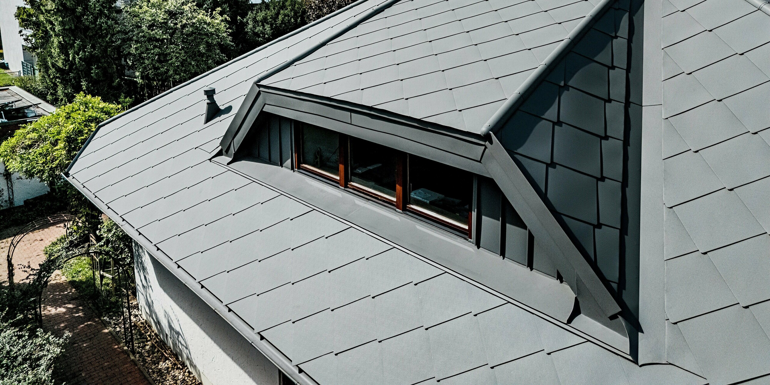 Das Bild zeigt eine eindrucksvolle Dachansicht eines Einfamilienhauses, das vollständig mit PREFA Dachrauten 44 × 44 in P.10 Hellgrau eingedeckt ist. Im Mittelpunkt steht die präzise ausgeführte Trapezgaube, deren klare Linien und glatte Aluminiumflächen harmonisch in die umliegende Rautenstruktur übergehen. Die hochwertige Stucco-Oberfläche verleiht dem Dach zusätzliche Tiefe und Stabilität. Unter einem lebhaften Himmel wirkt die langlebige, witterungsbeständige PREFA Dachlösung besonders elegant und modern, ideal für anspruchsvolle Sanierungsprojekte.