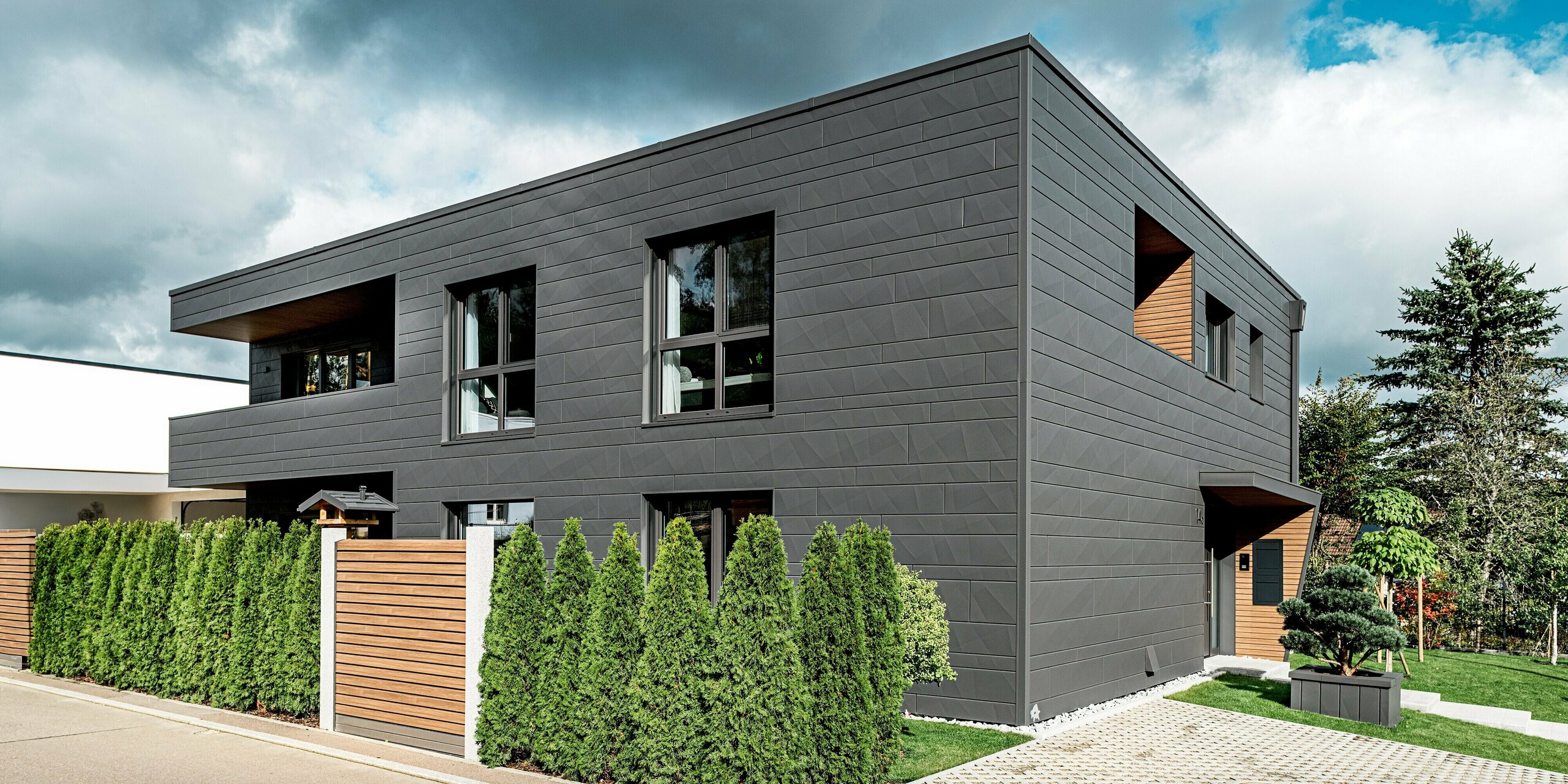 Moderne Seitenansicht eines Einfamilienhauses in Heidenheim mit vollständig verkleideter PREFA Siding.X Aluminiumfassade in P.10 Dunkelgrau. Die markanten Kantlinien und variierenden Paneelbreiten erzeugen eine dynamische Textur. Vor dem Gebäude zieht eine dichte Thujenhecke die Blickführung entlang der Grundstücksgrenze; ein Holzzaun setzt einen warmen Kontrast zur kühlen Metalloberfläche. Große Fensteröffnungen und holzverkleidete Rücksprünge verstärken den modernen Charakter. Die langlebige PREFA Oberfläche bietet hohen Witterungsschutz und ein präzises, zeitloses Fassadendesign.