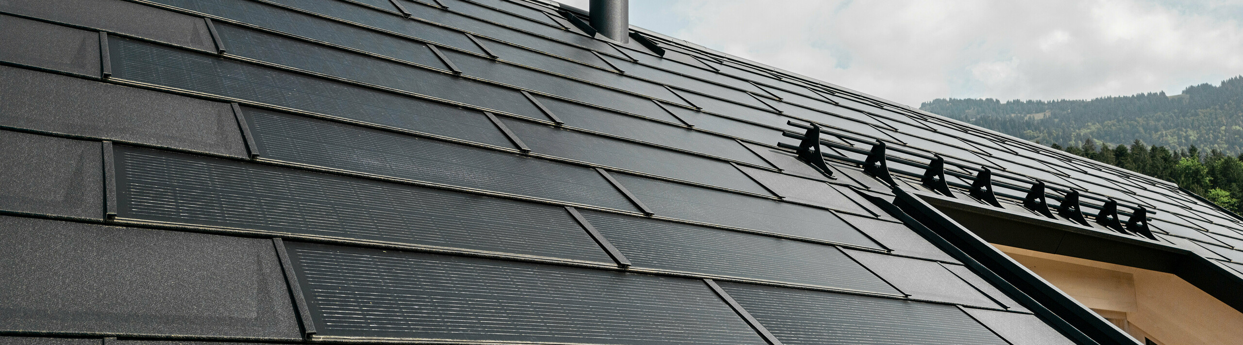 Nahaufnahme der effizienten PREFA Solardachplatte, welche sich nahtlos integriert in das schwarze Dach aus robusten PREFA Dachplatten R.16. Die Kombination aus nachhaltiger Energiegewinnung und langlebigem Aluminiumdach bietet eine ökologische und ästhetische Dachlösung, die sich harmonisch in die natürliche Landschaft einfügt.