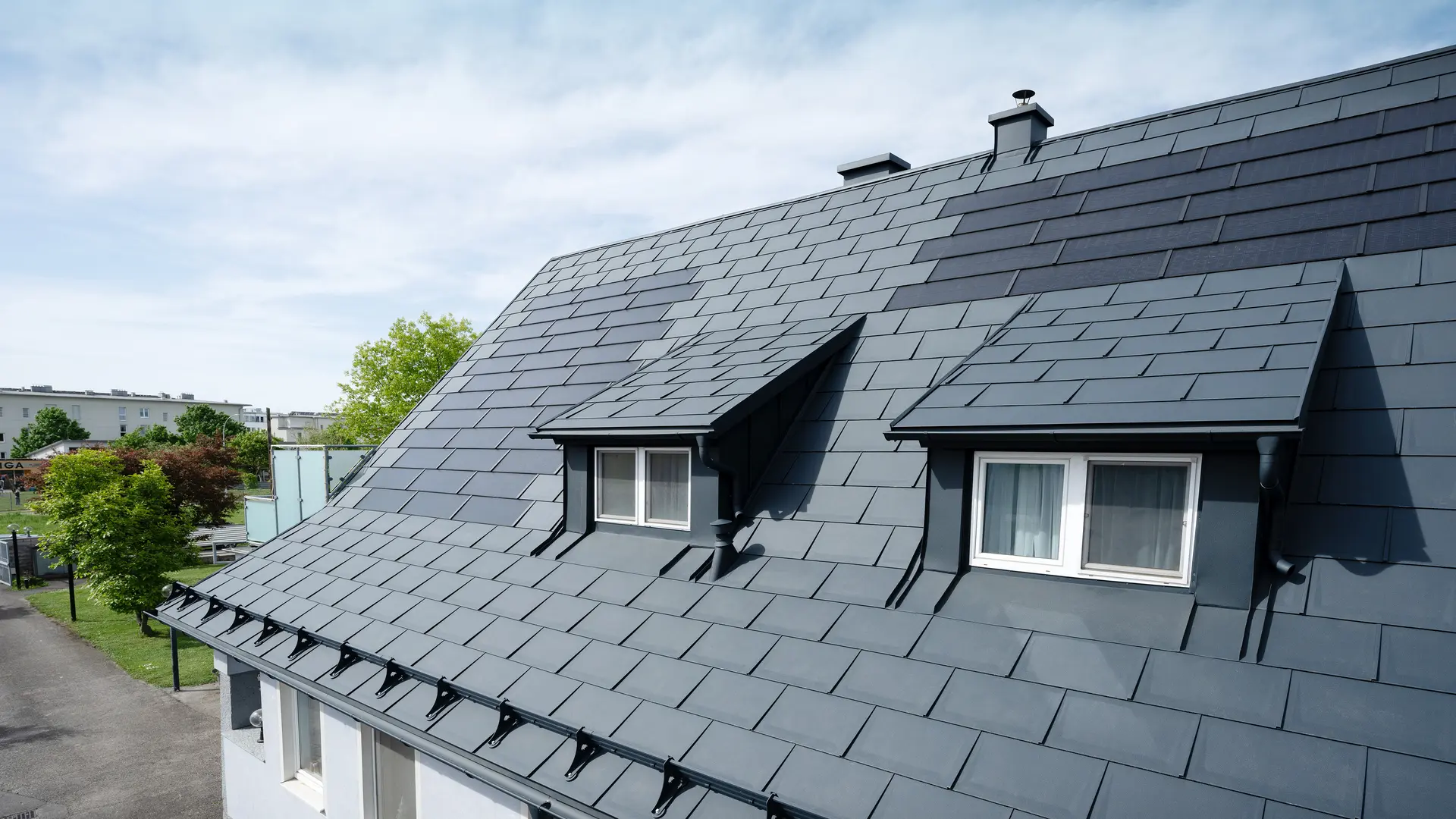 Detailaufnahme eines Dachs, das mit großen PREFA Solardachplatten und der PREFA Dachplatte R.16 in P.10 Anthrazit gedeckt ist. Zwei Gauben sind sichtbar. Die Solardachplatten sind gleichmäßig auf dem Dach verteilt und sorgen für eine moderne, nachhaltige Lösung.