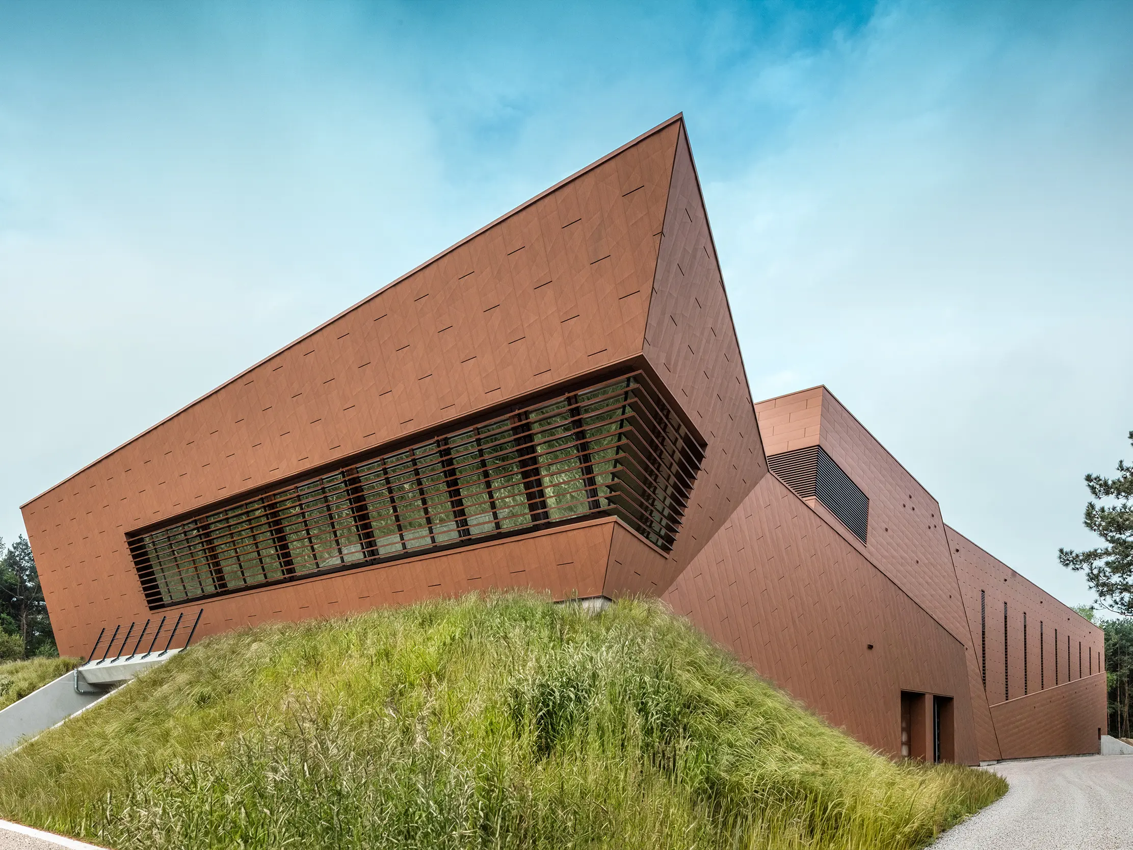 Markantes Trinkwasserpumpwerk von WML in Lottum mit skulptural geneigter Gebäudeform und prägnanter PREFA Siding.X Aluminiumfassade in warmem Rostton. Die dynamisch strukturierte Oberfläche reflektiert Licht und setzt starke architektonische Akzente im Landschaftsraum. Großzügige Lamellenöffnungen und klare Linienführung verbinden Technik und Gestaltung. Die langlebigen, leichten und korrosionsbeständigen PREFA Aluminiumpaneele überzeugen durch nachhaltige Recyclingfähigkeit, hohe und eine wartungsarme Gebäudehülle – ein moderner Eyecatcher für zukunftsorientierten Infrastrukturbau.