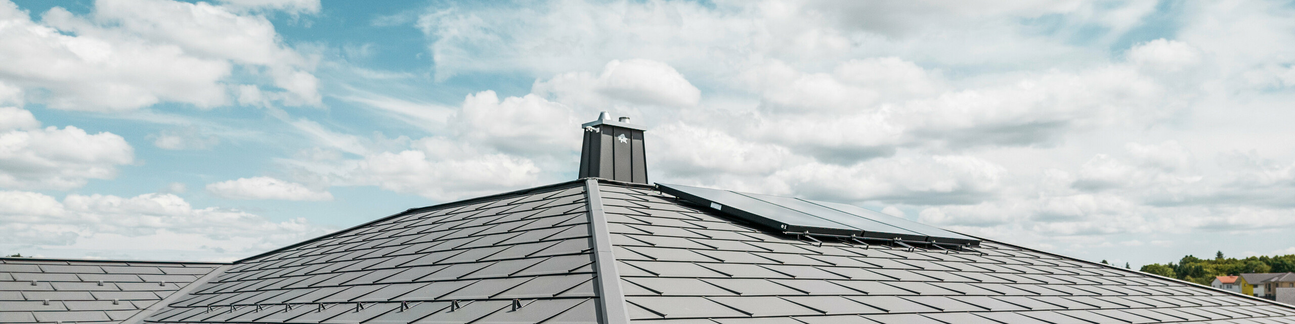 Detailansicht eines PREFA Aluminiumdachs auf einem Einfamilienhaus in Illmenau, Thürigen. Das Dach wurde überwiegend mit PREFA Dachplatten R.16 in P.10 Dunkelgrau eingedeckt. Die kleinformatigen Aluminiumdachplatten wurden fachmännisch verlegt und verleihen dem Bungalow einen modernen Look. Das Bild zeigt die Präzision des PREFA Dachsystems, inklusive der Schneefangsysteme, die für zusätzliche Sicherheit sorgen. Die harmonische Integration der Hängerinnen und Ablaufrohre des PREFA Dachentwässerungssystems betont die ästhetische und funktionale Qualität des Gebäudes. Am linken Bildrand sind Stehfalzstreifen zu sehen, welche mit dunkelgrauem PREFALZ auf der direkt anschließenden Garage montiert wurden.
