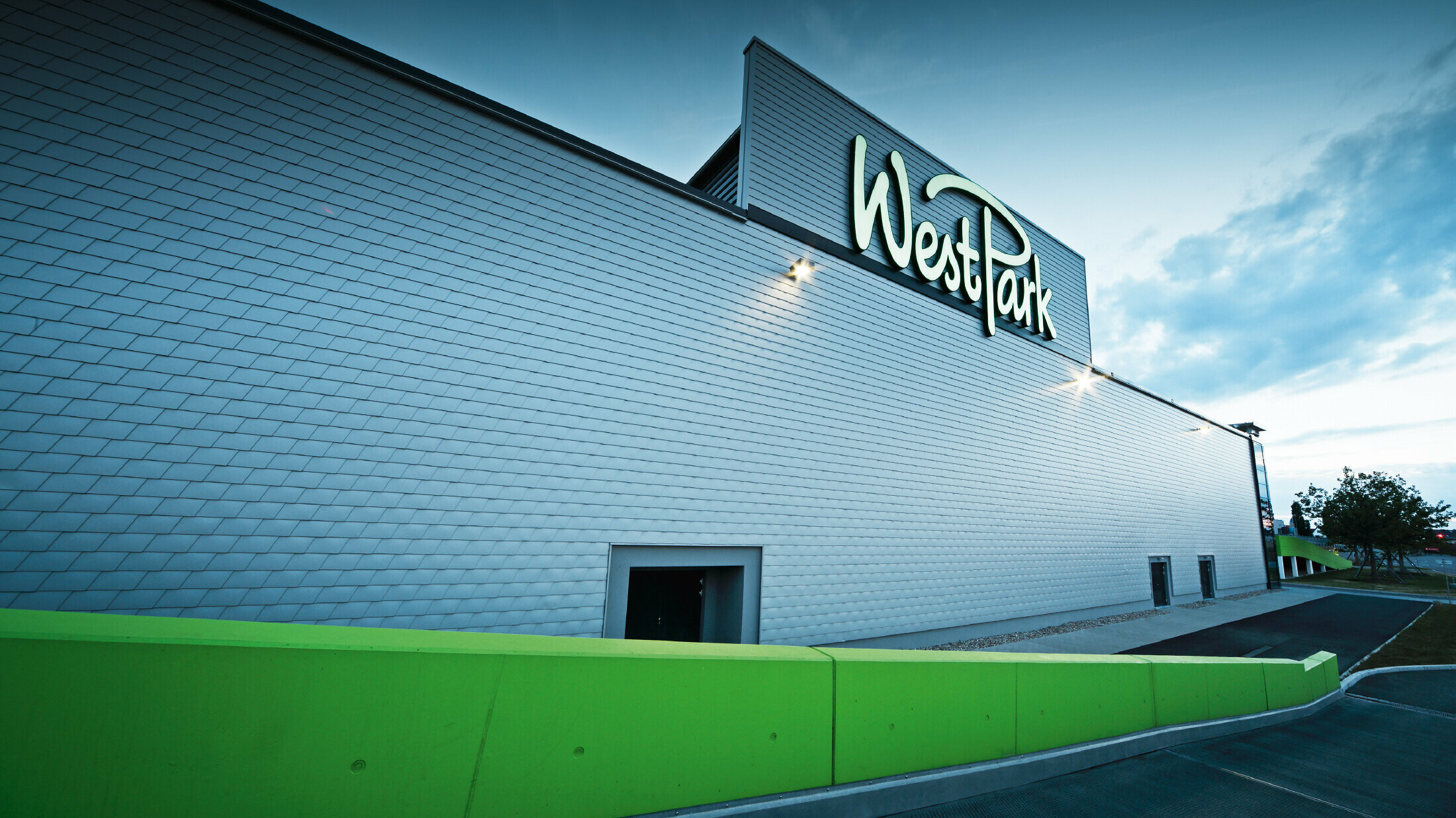 Moderne Fassade eines Einkaufszentrums mit großem Schriftzug „WestPark“. Die Außenwand ist mit silbermetallicfarbenen Aluminium-Schindeln verkleidet und wirkt durch die gleichmäßige Struktur elegant und technisch. Im Vordergrund verläuft eine leuchtend grüne Mauer entlang des Gehwegs. Das Gebäude ist in der Abenddämmerung beleuchtet.