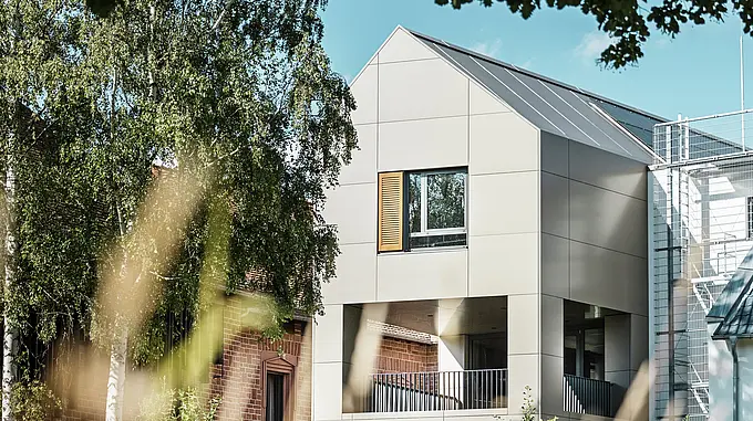 Das Bild zeigt das moderne Rathaus in Alzenau, dessen Fassade aus Aluminium-Verbundplatten von PREFA in einem eleganten Bronze-Ton besteht. Der spitze Dachbereich und die großen Fenster betonen den zeitgenössischen Charakter des Gebäudes. Im Vordergrund rahmen Bäume und Grünflächen das Gebäude ein, wobei die weiche Beleuchtung und der klare Himmel den modernen urbanen Raum unterstreichen.