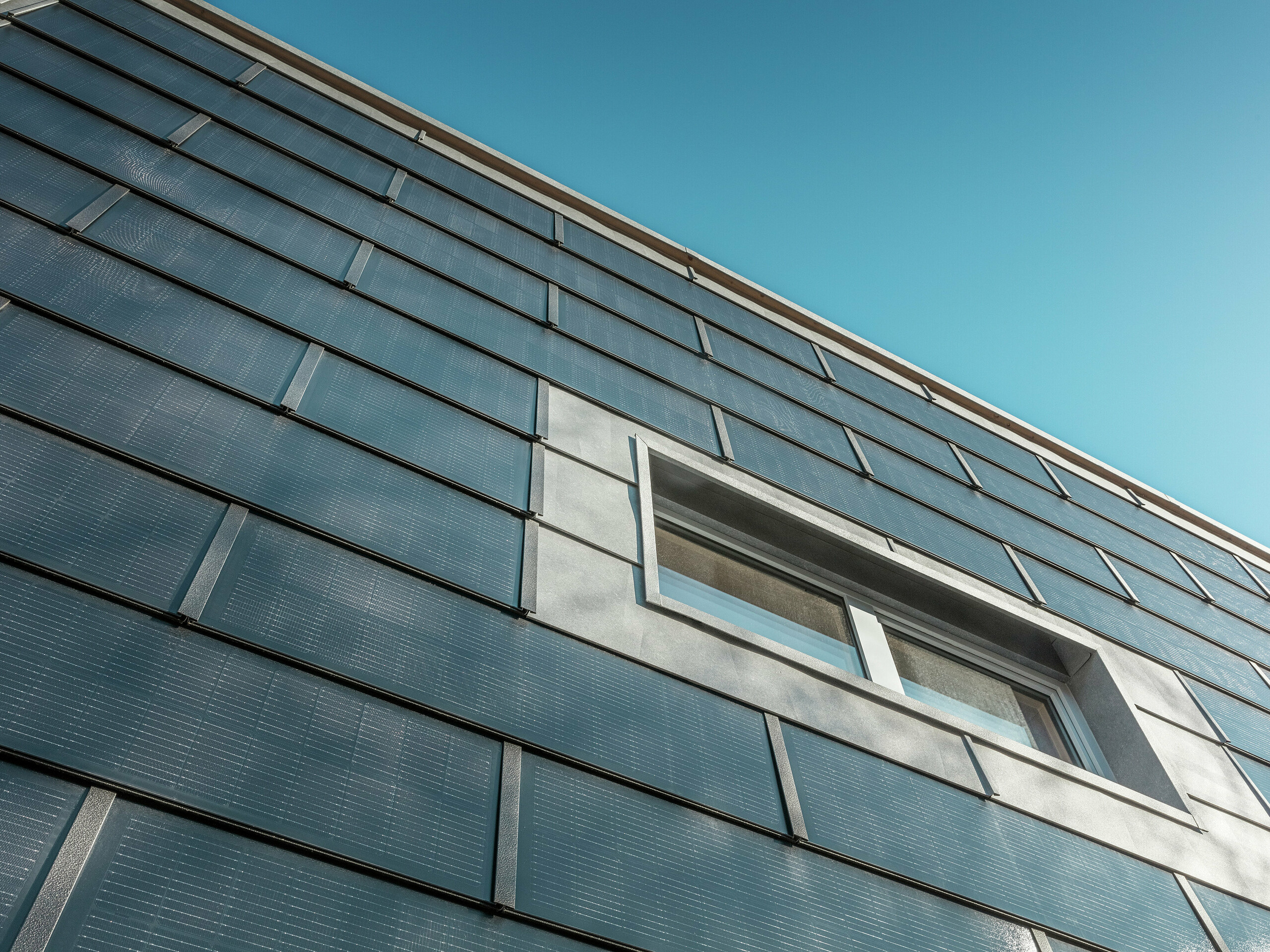 Die schräg aufgenommene Fassade der Spenglerei RÜMA in Vorarlberg ist mit PREFA Solardachplatten verkleidet. Die grauen Aluminiumelemente und die integrierten Photovoltaikmodule umrahmen ein Fenster. Im Hintergrund ist ein strahlend blauer Himmel zu erkennen.