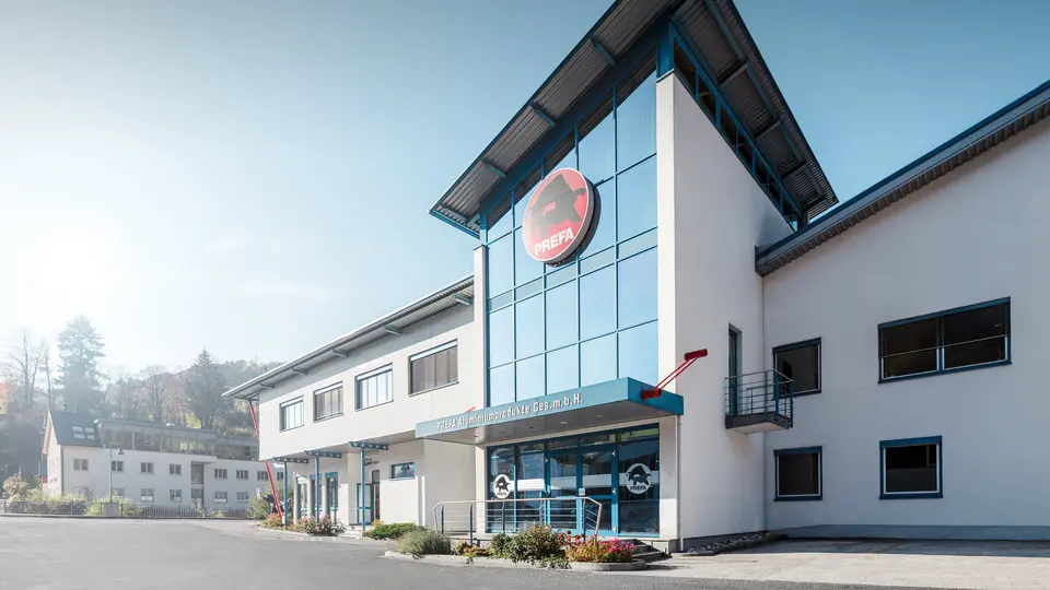 Spektakuläre Froschperspektive auf den PREFA Standort Marktl mit markantem PREFA-Stier-Logo, großzügiger Glasfassade und moderner Architektur. Der eindrucksvolle Eingangsbereich steht für Innovationskraft und Qualität. 