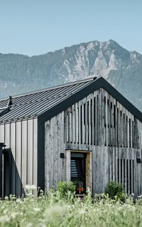Moderner, funktionaler Wohnbau mit einer Kombination aus Holzfassade und PREFA Band- und Blechmaterial für das Dach. Die klare Architektur wird durch die elegante, metallische Dach- und Fassadenverkleidung unterstrichen, die ideal für anspruchsvolle Projekte ist. Das Bild veranschaulicht die vielseitigen Anwendungsmöglichkeiten der PREFA Produkte im Bereich Bänder und Bleche.