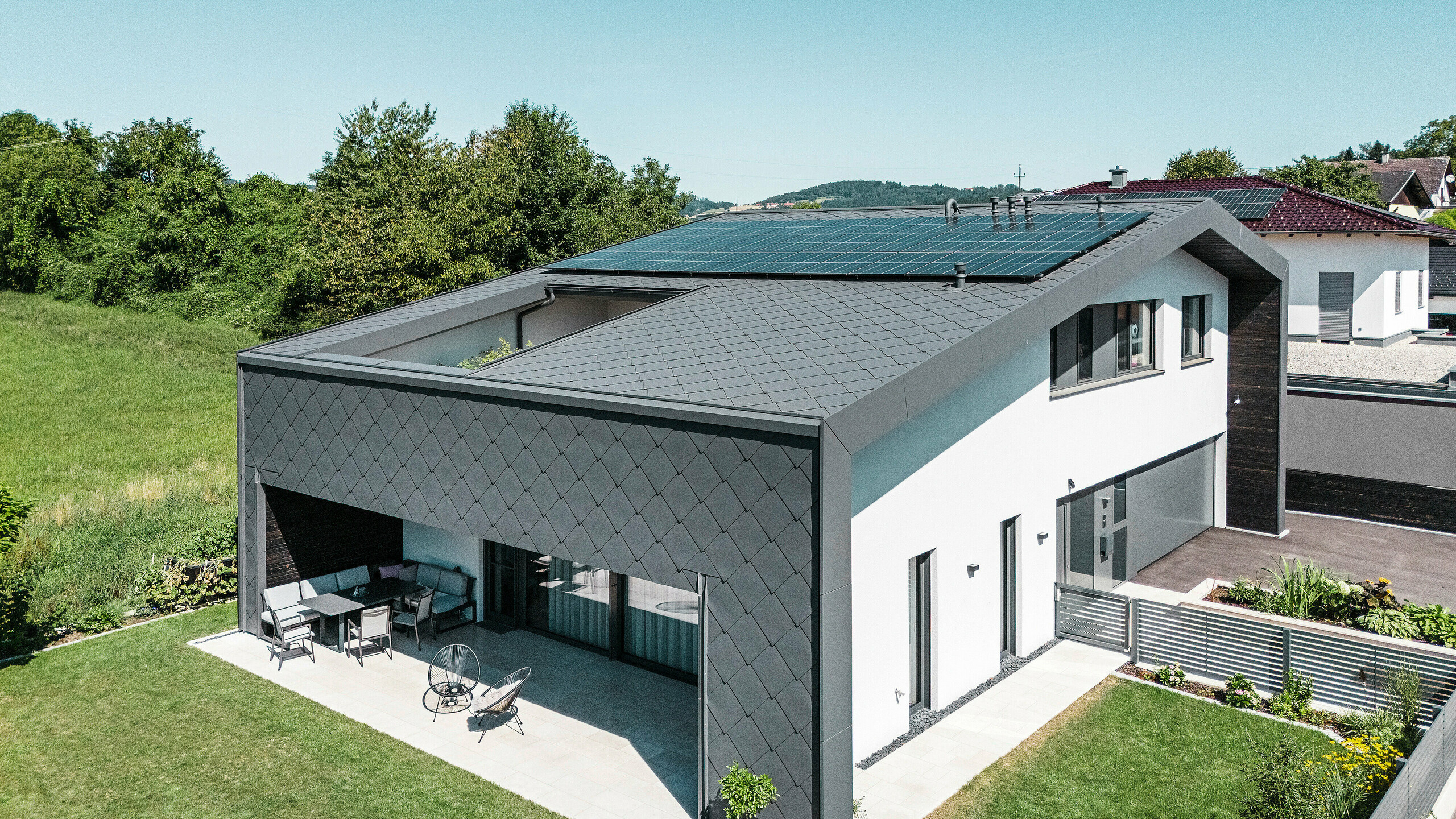 Modernes Einfamilienhaus in Aschach mit eleganter Aluminiumbekleidung aus dunkelgrauen PREFA Dach- und Wandrauten 44 × 44 in der hochwertigen Beschichtung P.10. Das Gebäude zeichnet sich durch eine großzügige, überdachte Terrasse aus, die fließend in den gepflegten Garten übergeht. Eine Photovoltaikanlage auf dem Dach sorgt für nachhaltige Energiegewinnung. Die Kombination aus moderner Architektur, witterungsbeständiger Aluminiumhülle und schlichter weißer Putzfassade verleiht dem Haus ein stilvolles und zeitgemäßes Erscheinungsbild.