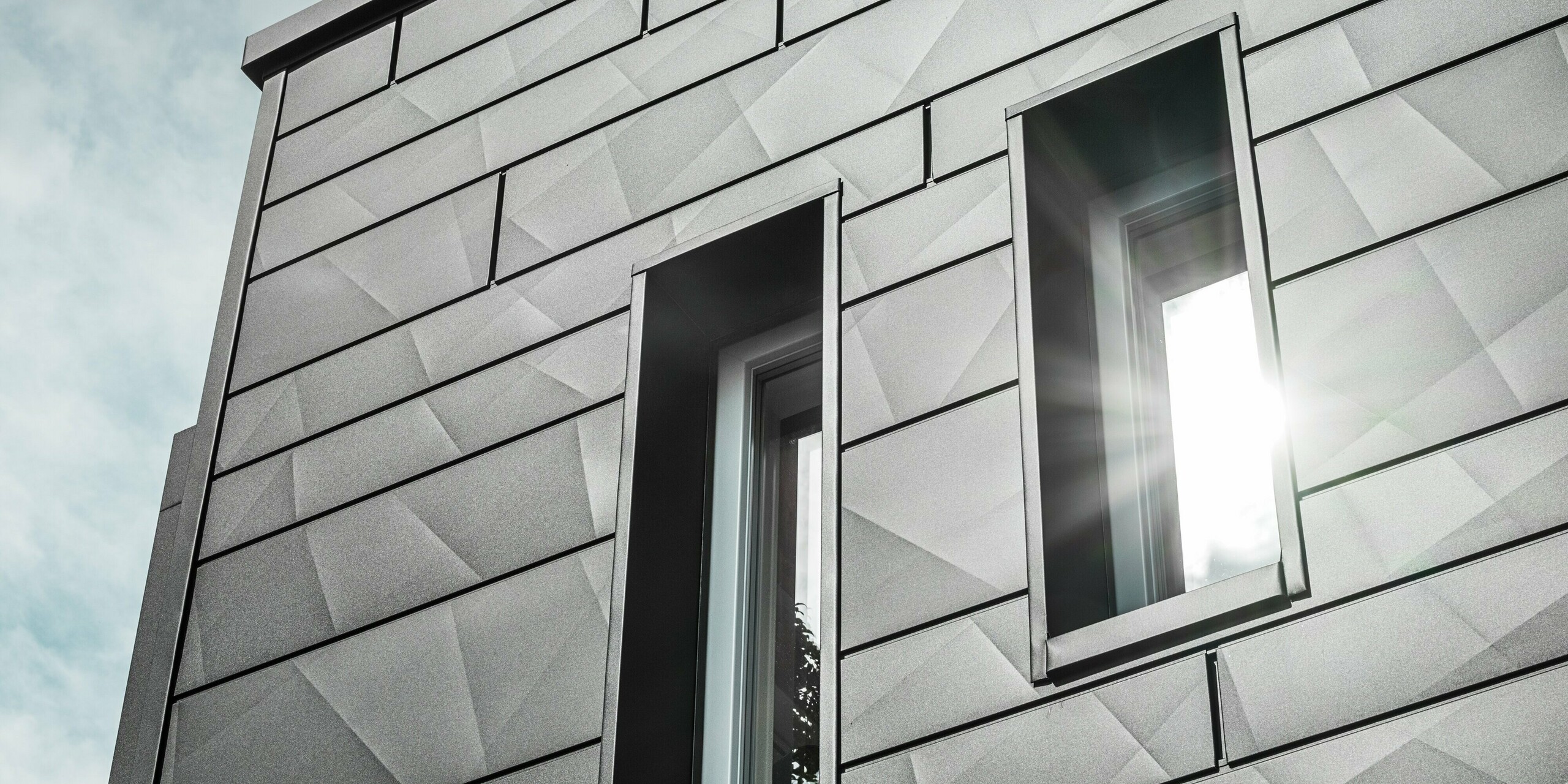 Die moderne Fassade des sanierten Einfamilienhauses in Teuschnitz zeigt die geometrischen PREFA Siding.X Fassadenpaneele in P.10 Schwarz. Die Fensterumrahmungen aus schwarzen Aluminium Verbundplatten fügen sich harmonisch in das Gesamtbild ein. Ein Sonnenstrahl fällt durch eines der Fenster und betont die klare, strukturierte Oberfläche der Fassadenpaneele. Die ästhetische Wirkung der Fassade wird durch das subtile Zusammenspiel von Licht und Material verstärkt.