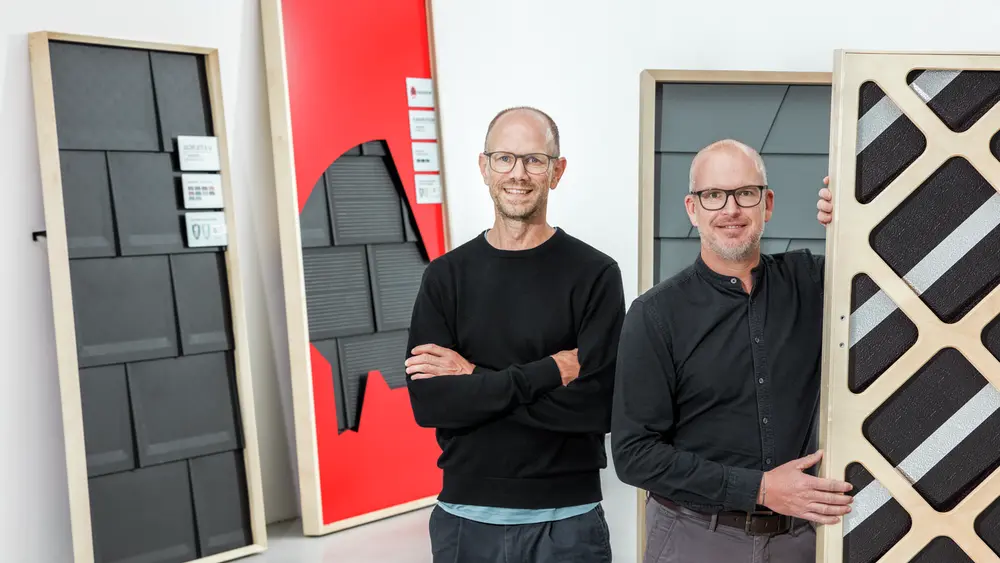 Der Kreativdirektor Markus Vogler von AERA und Marketingleiter Jürgen Jungmair von PREFA lächeln in die Kamera. Im Hintergrund sieht man das neue Messekonzept von PREFA.