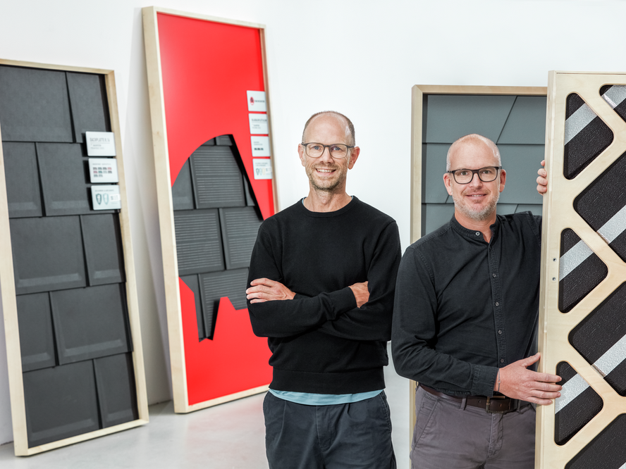 Der Kreativdirektor Markus Vogler von AERA und Marketingleiter Jürgen Jungmair von PREFA lächeln in die Kamera. Im Hintergrund sieht man das neue Messekonzept von PREFA.
