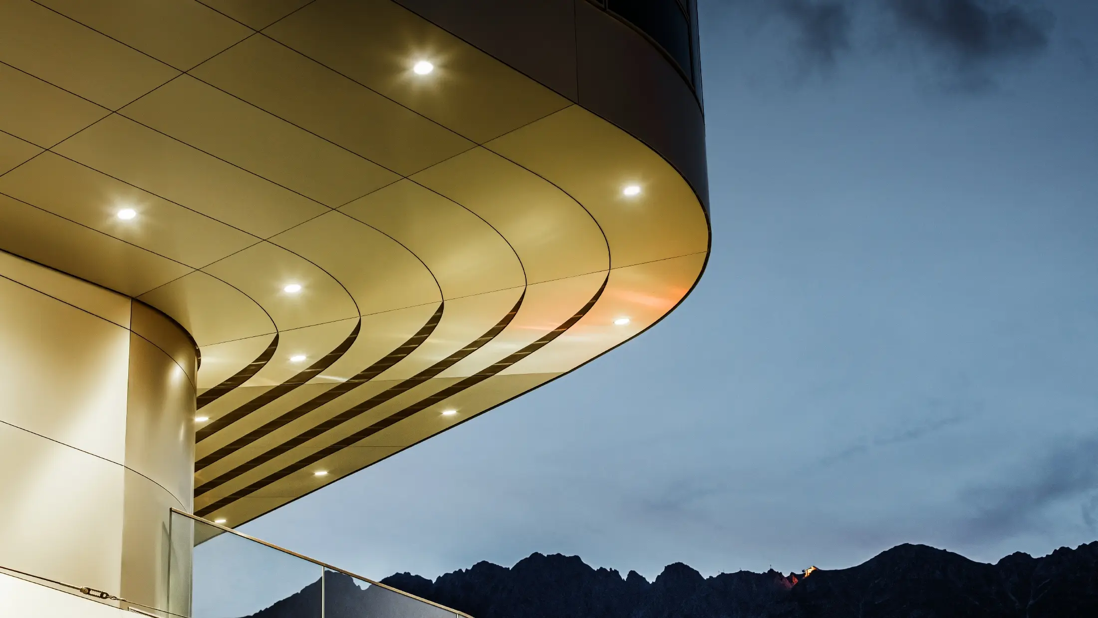 Das Bild zeigt eine beeindruckende architektonische Detailsansicht einer PREFA PEMA Verbundplatte aus gebürstetem Aluminium. Die Platte ist sanft gewölbt und verläuft in eleganten, fließenden Linien, die durch gezielte Beleuchtung am Abend besonders zur Geltung kommen. Die klare Struktur und der edle Aluminium-Look der Platte ergänzen das moderne Design der Fassade und bieten eine elegante, zeitgenössische Lösung für hochwertige Architektur.