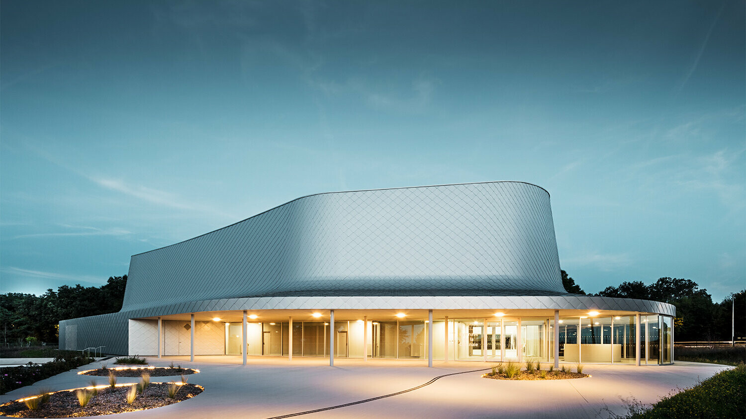 Seitliche Abendaufnahme der von Bohuon Bertic Architectes entworfenen Sporthalle in Basse-Goulaine mit Beleuchtung.