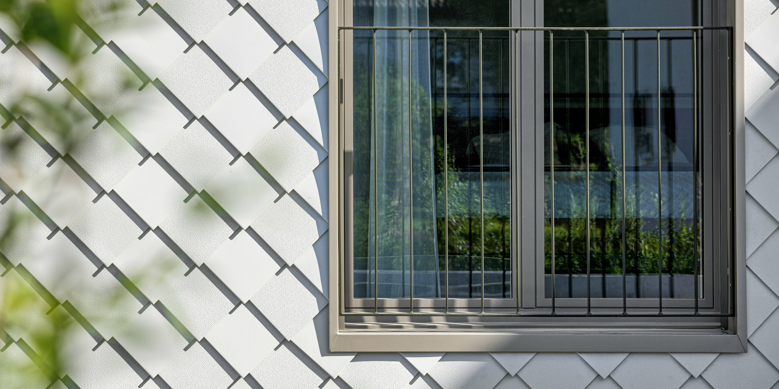 Architektonisch anspruchsvolle Fassade eines modernen Wohnhauses mit PREFA Wandrauten 20x20 in der Farbe P.10 Prefaweíß. Die langlebigen und wartungsarmen Rauten aus Aluminium umrahmen ein Fenster mit einem französischen Balkon. Die Verkleidung bietet ein leichtes, aber robustes Fassadensystem, das dem Objekt ein ästhetisch reizvolles, skaliertes Erscheinungsbild verleiht.