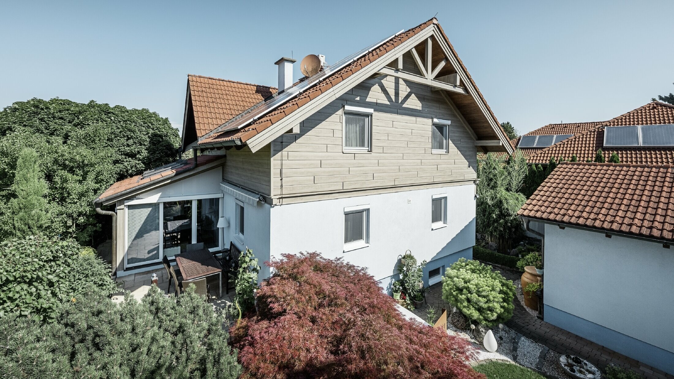 Einfamilienhaus mit Satteldach und roter Dacheindeckung, kombiniert mit einer modernen Fassadengestaltung. Der obere Giebel ist mit beige-grauen Aluminium-Fassadenpaneelen von PREFA verkleidet, während das Erdgeschoss eine weiße Putzfassade hat. Auf dem Dach befinden sich Solarmodule. Das Haus liegt in einem grünen Garten mit Terrasse, Gartenmöbeln und üppiger Bepflanzung, umgeben von weiteren Wohnhäusern mit ähnlicher Dacharchitektur.