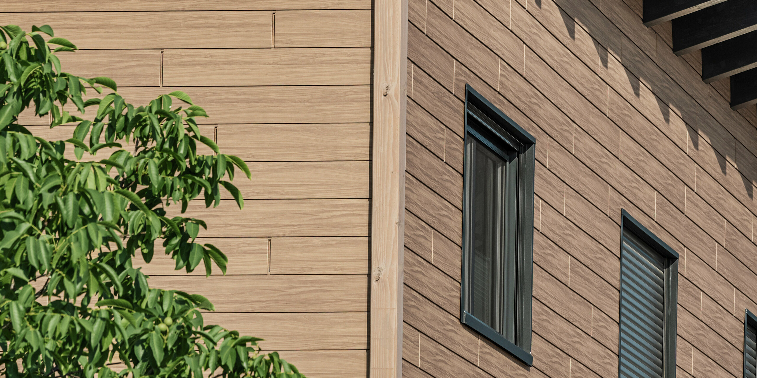 Blick von unten auf die Gebäudekante am Obergeschoss eines Einfamilienhauses in Bruck. Das obere Stockwerk ist mit PREFA Sidings in Walnuss Braun verkleidet. Die Aluminiumfassade in Holzoptik sorgt für einen harmonischen Gesamteindruck des Gebäudes.