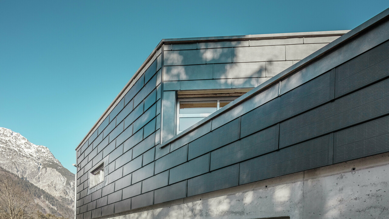 Die Fassade des Bürogebäudes der RÜMA Blechwerkstatt in Vorarlberg mit innovativen PREFA Solardachpaneelen und Sichtbeton, umgeben von Bergen unter klarem blauem Himmel, mit Baumschatten an der Wand.