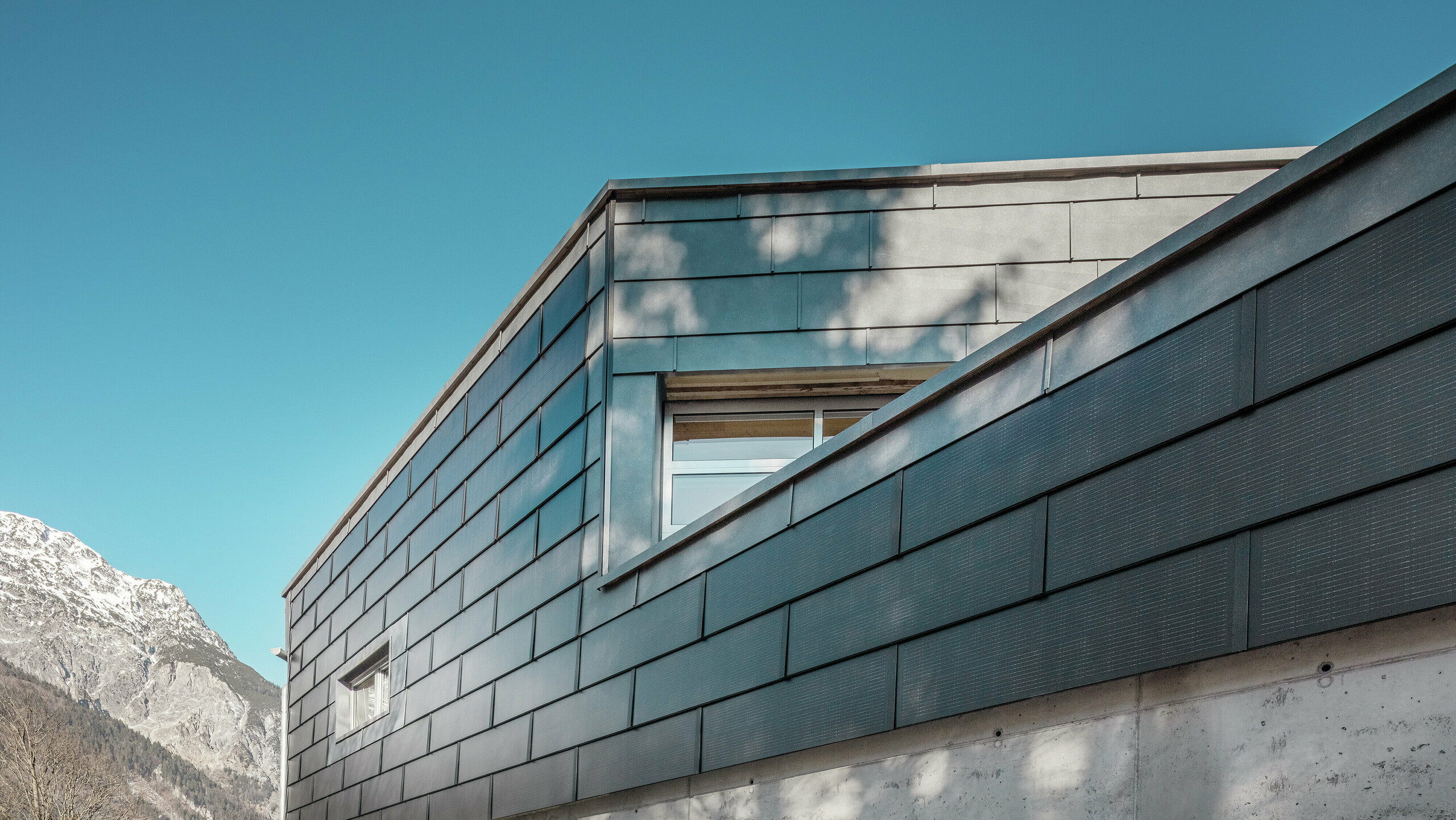 Fassade des Firmengebäudes der Spenglerei RÜMA in Vorarlberg mit innovativen PREFA Solardachplatten und Sichtbeton, umgeben von Bergen unter klarem blauem Himmel, mit Schatten von Bäumen auf der Wand.