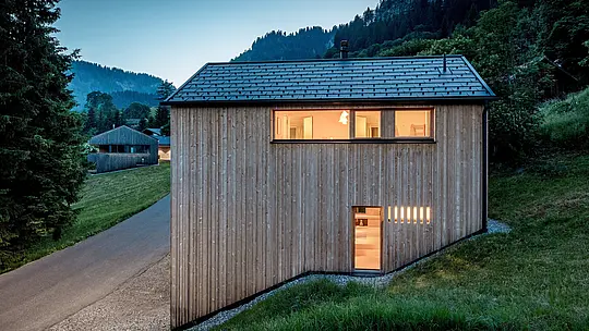 Vor der dramatischen Bergkulisse von Ebnit wirkt Haus Immi wie ein warmer Rückzugsort im Wald. Die beleuchteten Fenster betonen die schlichte Weißtannenfassade, während das schwarze PREFA Solardach die klare Silhouette prägt. Integrierte Solardachplatten und Dachplatten R.16 in P.10 Schwarz erzeugen nachhaltig Strom und bieten robusten, langlebigen Wetterschutz im alpinen Raum.