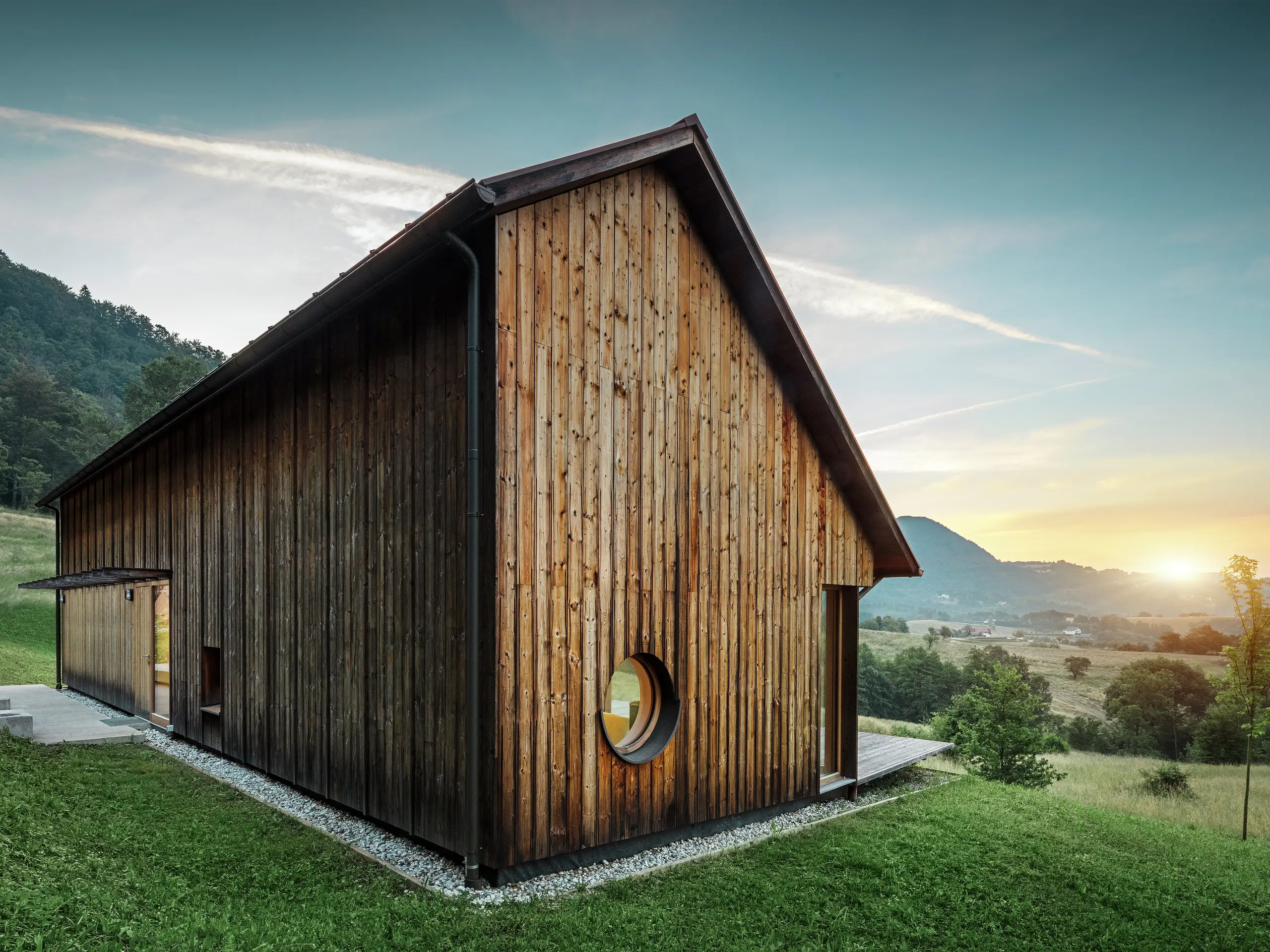 Dieses beeindruckende Foto zeigt ein Holzhaus in Vinje im Sonnenuntergang mit einem robusten PREFALZ-Aluminiumdach in der Farbe P.10 Braun. Das witterungsbeständige Stehfalzdach von PREFA ist langlebig, wartungsarm und fügt sich harmonisch in die Landschaft ein. Die Kombination aus klaren Linien, Holzfassade und Rundfenster unterstreicht den architektonischen Minimalismus. Das schlichte Satteldach besticht durch seine bewusst asymmetrische Form, die ihm einen besonderen Charakter verleiht.