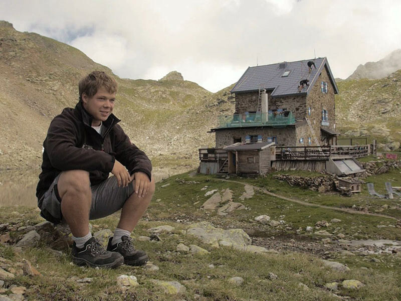 Lehrling Jakob Schöttel aus Bayern sitzt vor der Flagger Schartenhütte in Südtirol und erzählt über seine Arbeit. Im Hintergrund sieht man die Hütte mit der PREFA Dachplatte in Steingrau eingedeckt.