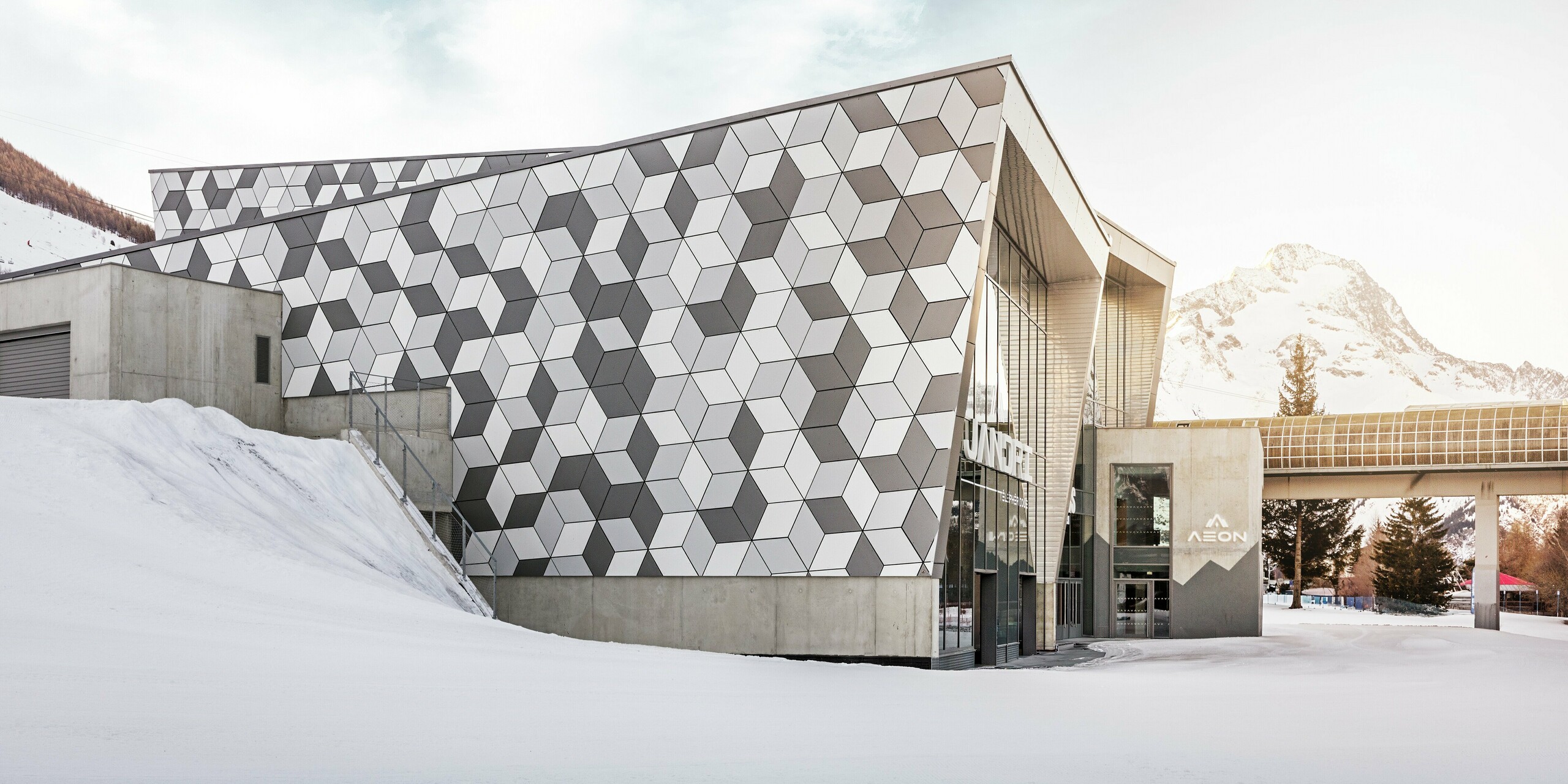 Die Talstation in Les Deux Alpes beeindruckt mit einer außergewöhnlichen Fassade aus PREFABOND Aluminium-Verbundplatten in einem modernen Rautenmuster. Die Kombination aus Weiß- und Grautönen bildet ein markantes Design, das perfekt mit der verschneiten Berglandschaft harmoniert. Die langlebige, wetterbeständige Fassade garantiert auch unter extremen Bergbedingungen eine ästhetische und funktionale Lösung. Die strahlende Sonne im Hintergrund schafft einen majestätischen Rahmen für diese Szenerie.