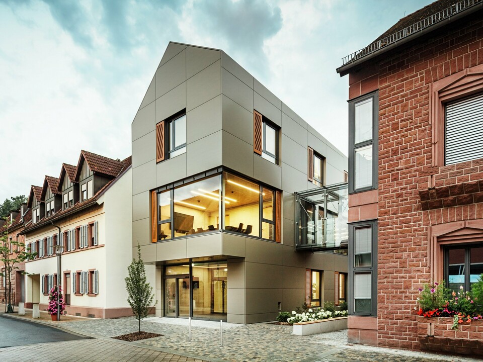 Das Bild zeigt das moderne Rathaus in Alzenau, das mit Aluminium-Verbundplatten von PREFA in einem warmen Bronze-Ton verkleidet ist. Die Fassade wird durch große Fensterflächen unterbrochen, die einen Blick auf den Innenraum gewähren, der durch warmes Licht erleuchtet wird. Der gläserne Verbindungsgang im rechten Teil des Bildes verbindet das Gebäude mit einem benachbarten, traditionellen Gebäude aus Ziegelstein, was einen spannenden Kontrast zwischen moderner und historischer Architektur schafft. 