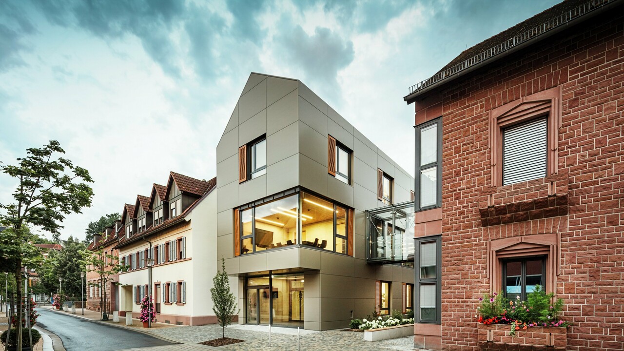 Das Bild zeigt das moderne Rathaus in Alzenau, dessen Fassade mit PREFA-Aluminiumverbundplatten in einem warmen Bronzeton verkleidet ist. Große Glasflächen unterbrechen die Fassade und geben den Blick auf ein lichtdurchflutetes Interieur frei. Ein gläserner Verbindungsgang auf der rechten Bildseite verbindet das Gebäude mit einem nahegelegenen traditionellen Backsteingebäude und schafft so einen interessanten Kontrast zwischen moderner und historischer Architektur.