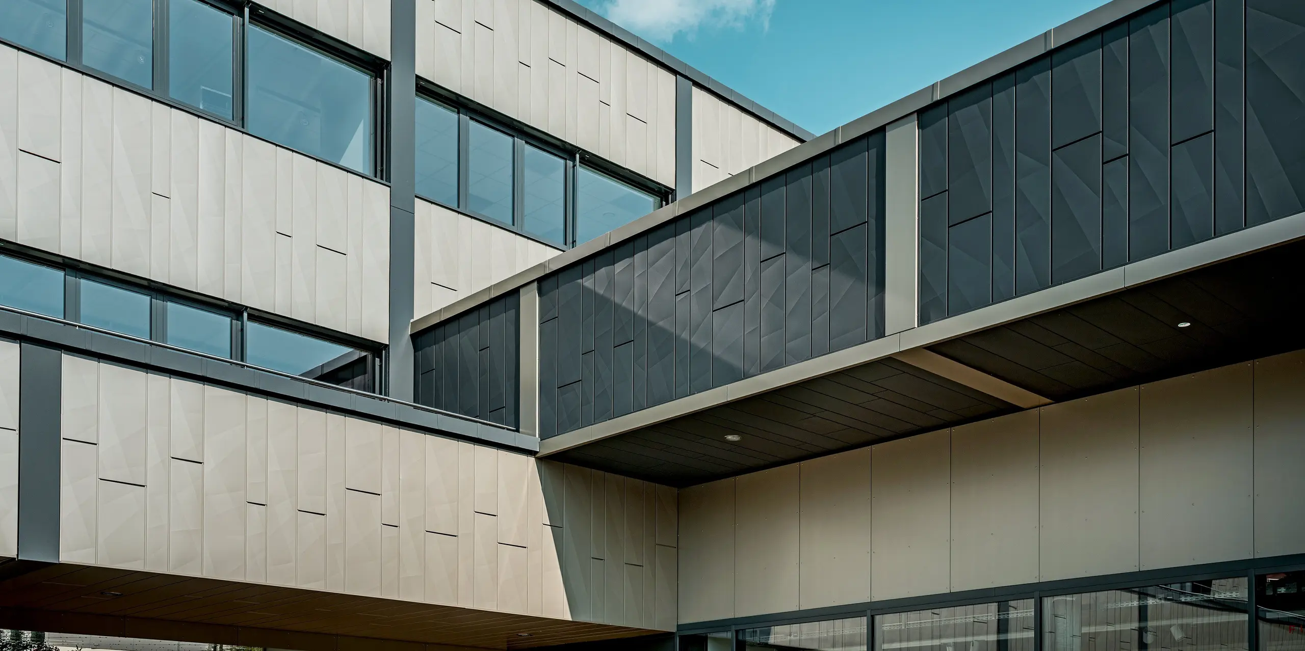 Die beeindruckende Aluminiumfassade des Einkaufszentrums in Rohrbach wurde mit PREFA Siding X-Fassadenpaneelen in den Farben Bronze und Anthrazit ausgestattet. Besonders auffällig ist der elegante Übergang zwischen den beiden Farben, der der Fassade eine dynamische und moderne Ausstrahlung verleiht. Die klare Struktur der Siding.X-Elemente sorgt für eine markante Gliederung der Wandflächen. Im Fokus stehen insbesondere die geometrischen Muster der Platten, die für eine besondere Tiefe sorgen.