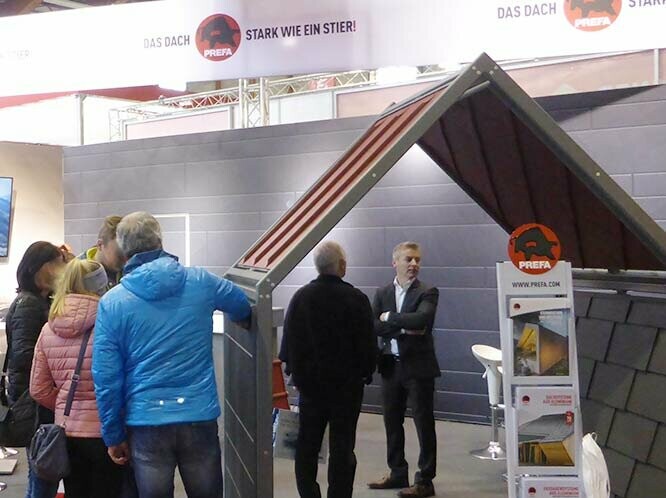 PREFA Messestand auf der Häuslbauermesse Klagenfurt 2018, zwei kleine Musterhäuser präsentieren die sturmsicheren Aluminiumprodukte für Dach und Fassade von PREFA, davor sind einige Personen zu erkennen