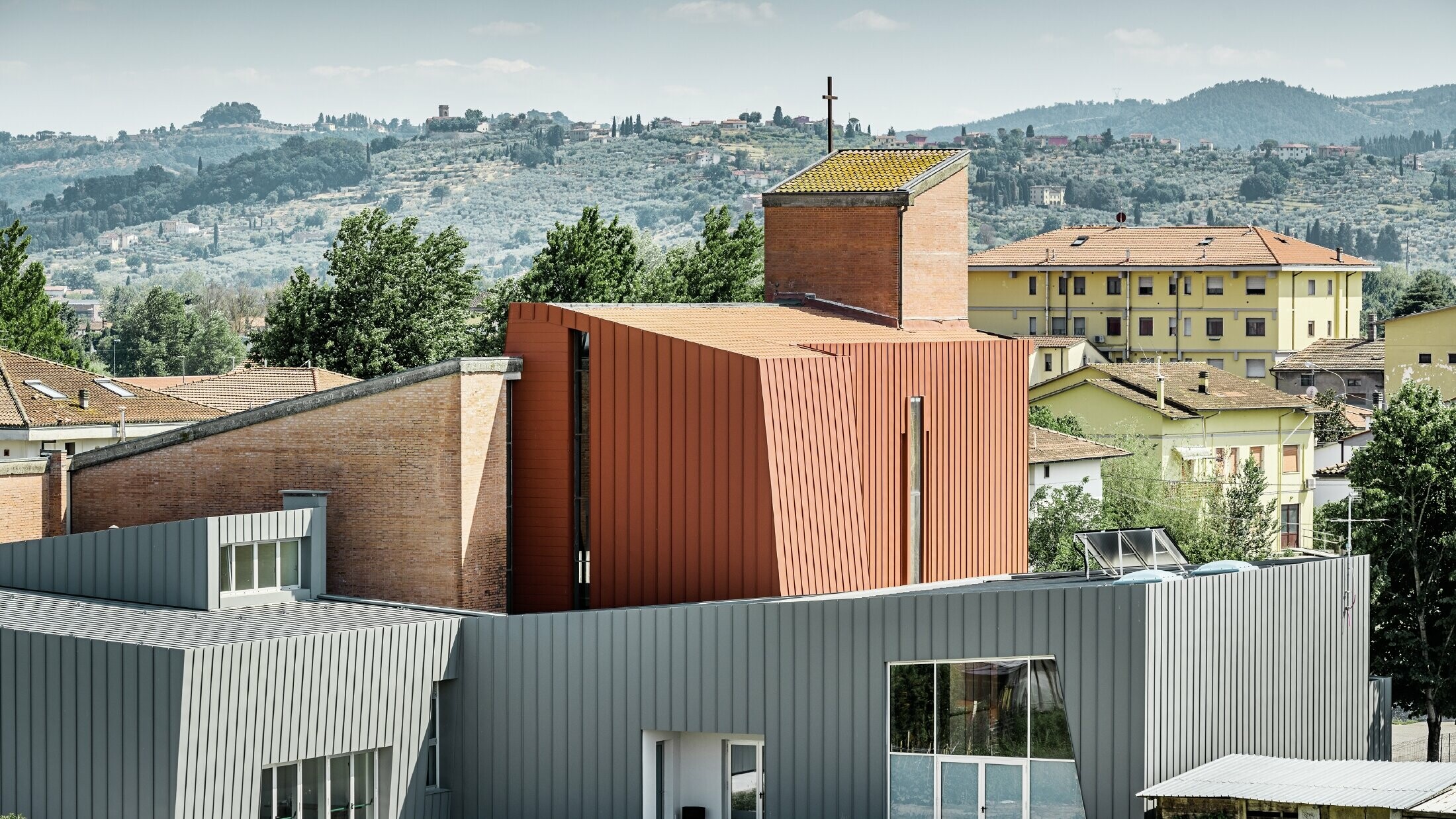 Modernes Kirchengebäude mit markanter Architektur, bestehend aus klaren geometrischen Formen. Der Bau kombiniert hellgraue Aluminiumfassaden mit einem zentralen, ziegelroten Turm aus Metallpaneelen und Ziegelmauerwerk, auf dessen Spitze ein Kreuz steht. Im Hintergrund sind Wohnhäuser und eine hügelige, grüne Landschaft mit Bäumen und Olivenhainen zu sehen.