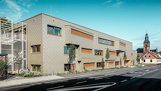 Bei strahlendem Tageslicht zeigt sich die moderne Kindertagesstätte in Alzenau mit ihrer prägnanten PREFA Aluminiumfassade aus bronzefarbenen Wandrauten 44 × 44 in voller Klarheit. Die gleichmäßige Schuppenstruktur, unterbrochen von versetzten Fenstern und vertikalen Holzlamellen, verleiht der Fassade Tiefe und Rhythmus. Links im Bild ist das filigrane Treppenhaus mit Metallgitterstruktur sichtbar, das für Transparenz und Leichtigkeit sorgt. Im Hintergrund steht die spätgotische Kirche von Alzenau – Symbol für die harmonische Verbindung von historischer Baukultur und zeitgemäßer Architektur. Das wartungsfreie, langlebige PREFA Fassadensystem unterstützt die nachhaltige, funktionale und ästhetisch hochwertige Gestaltung dieses zukunftsweisenden Bildungsbaus.