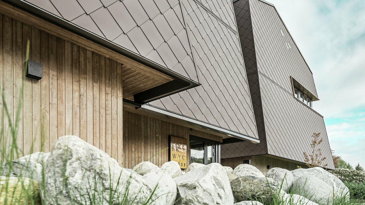 Seitliche Ansicht eines Hotels, die Fassade des Erdgeschosses ist mit eher dunklem Holz vertikal verkleidet. Darüber ist ein Fassadenvorsprung, ab hier ist die Fassade mit Aluminiumplatten in Nussbraun verkleidet.