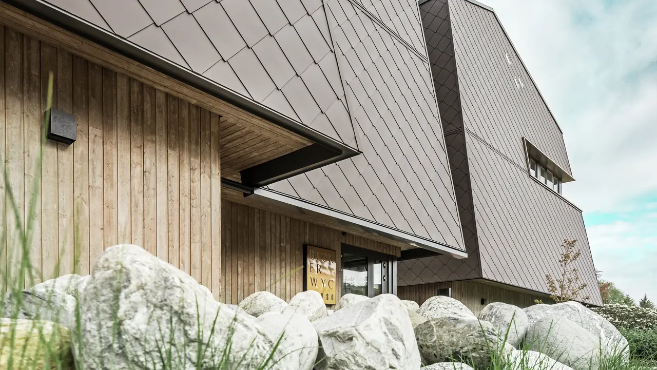 Seitliche Ansicht eines Hotels, die Fassade des Erdgeschosses ist mit eher dunklem Holz vertikal verkleidet. Darüber ist ein Fassadenvorsprung, ab hier ist die Fassade mit Aluminiumplatten in Nussbraun verkleidet.