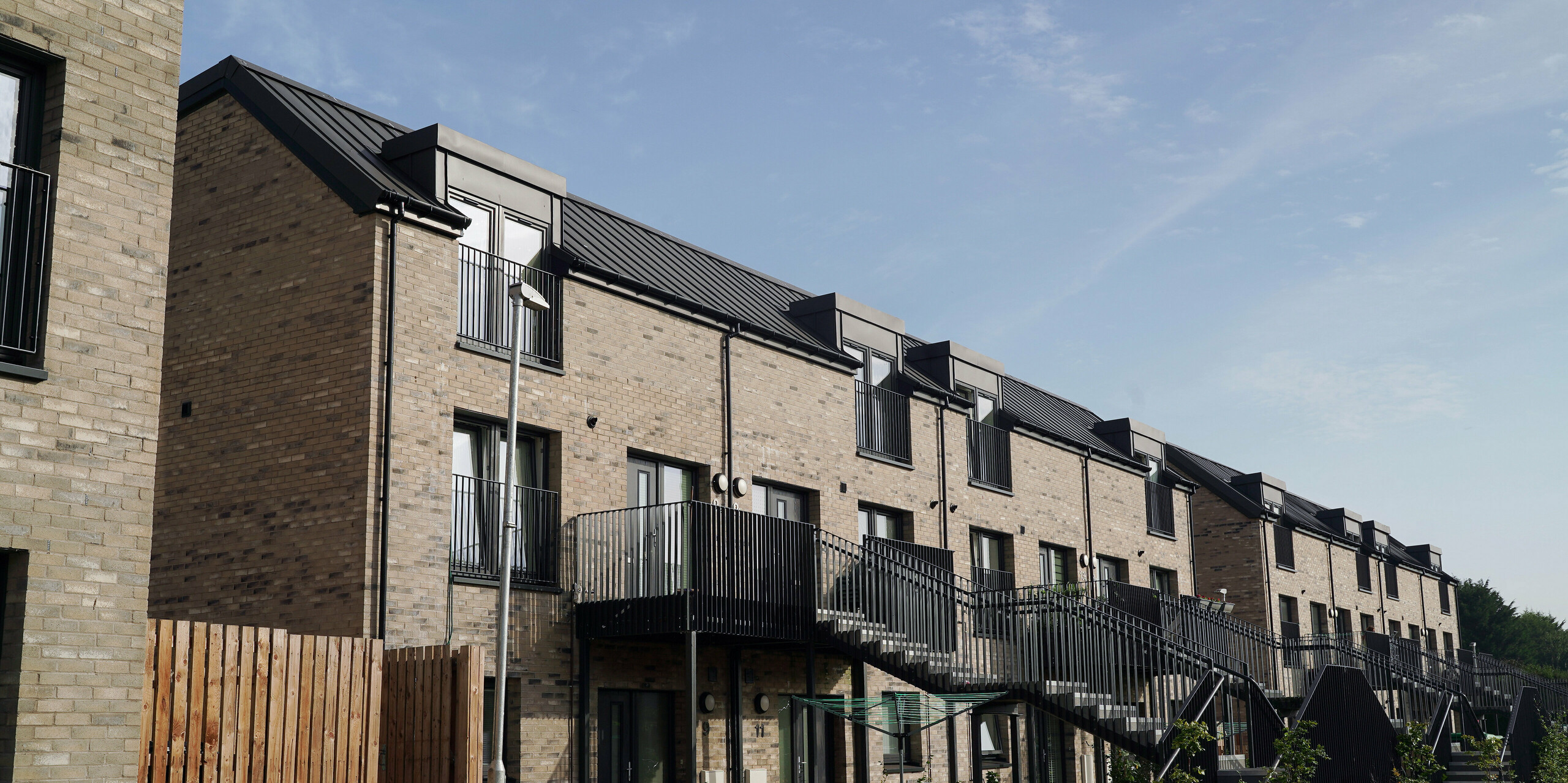 Nahaufnahme einer neu gebauten Wohnhausanlage in der Bingham Avenue, Edinburgh, mit PREFALZ Dächern in P.10 Dunkelgrau. Die dunklen Aluminiumdächer bieten einen ansprechenden Kontrast zu den hellen Ziegelmauern der Gebäude. Die einzelnen Wohnungen verfügen über einen Balkon bzw. Vorgarten. Die Stiegen sind mit schwarzen Geländern ausgestattet, die ein modernes und stilvolles Erscheinungsbild vermitteln. Die Vordergärten sind mit jungen Bäumen bepflanzt, was zur ästhetischen und ökologischen Qualität des Wohnkomplexes beiträgt.