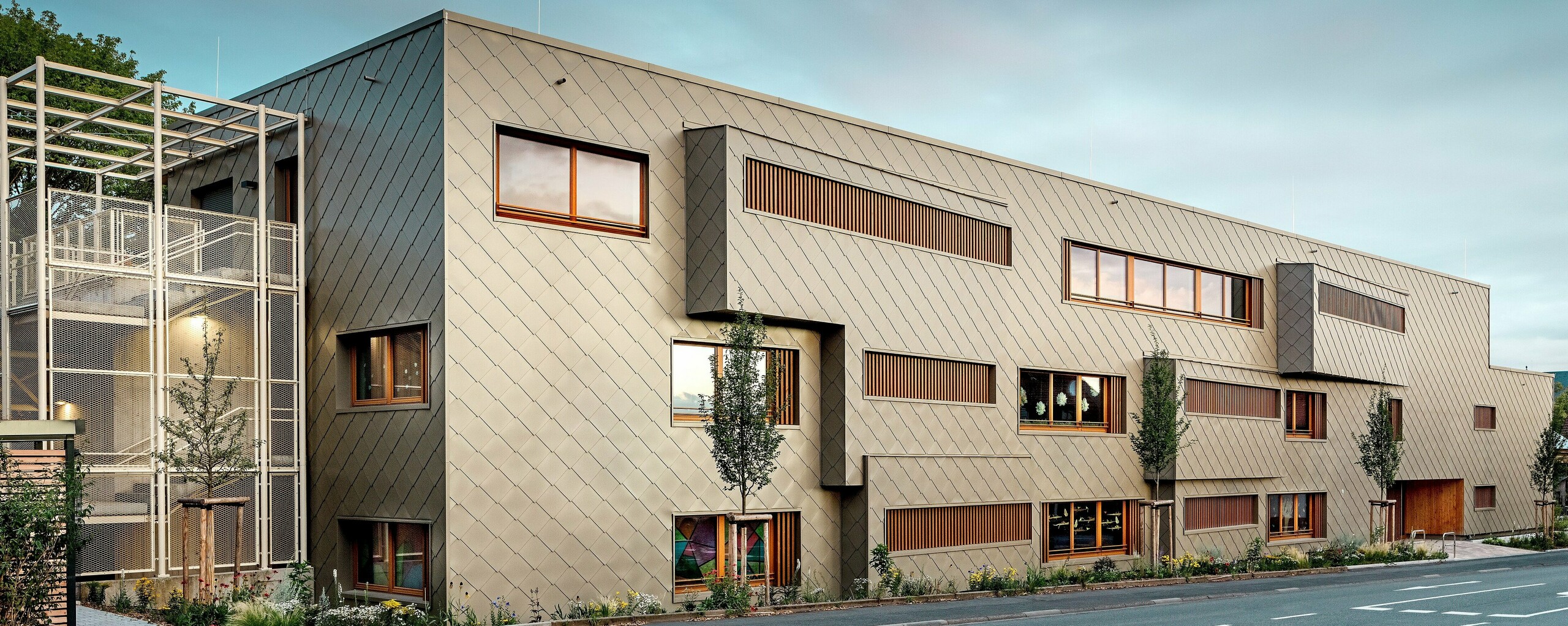 Die Aufnahme aus Straßenniveau zeigt die neue Kindertagesstätte in Alzenau im spannungsvollen Dialog mit der historischen Stadtkulisse, darunter die markante Kirche im Hintergrund. Die moderne Fassade aus bronzefarbenen PREFA Wandrauten 44 × 44 verleiht dem Gebäude eine elegante, fast skulpturale Wirkung. Präzise gesetzte Fenster, vertikale Holzlamellen und versetzte Fassadenflächen schaffen Tiefe und Rhythmus. Links ist das filigrane Treppenhaus mit weißem Metallgerüst sichtbar, das moderne Leichtigkeit vermittelt. Die klare Formsprache, nachhaltige Materialwahl und die hochwertige Aluminiumfassade machen das Gebäude zu einem Vorzeigeprojekt für städtebauliche Integration und zukunftsorientiertes Bauen mit PREFA Systemen.