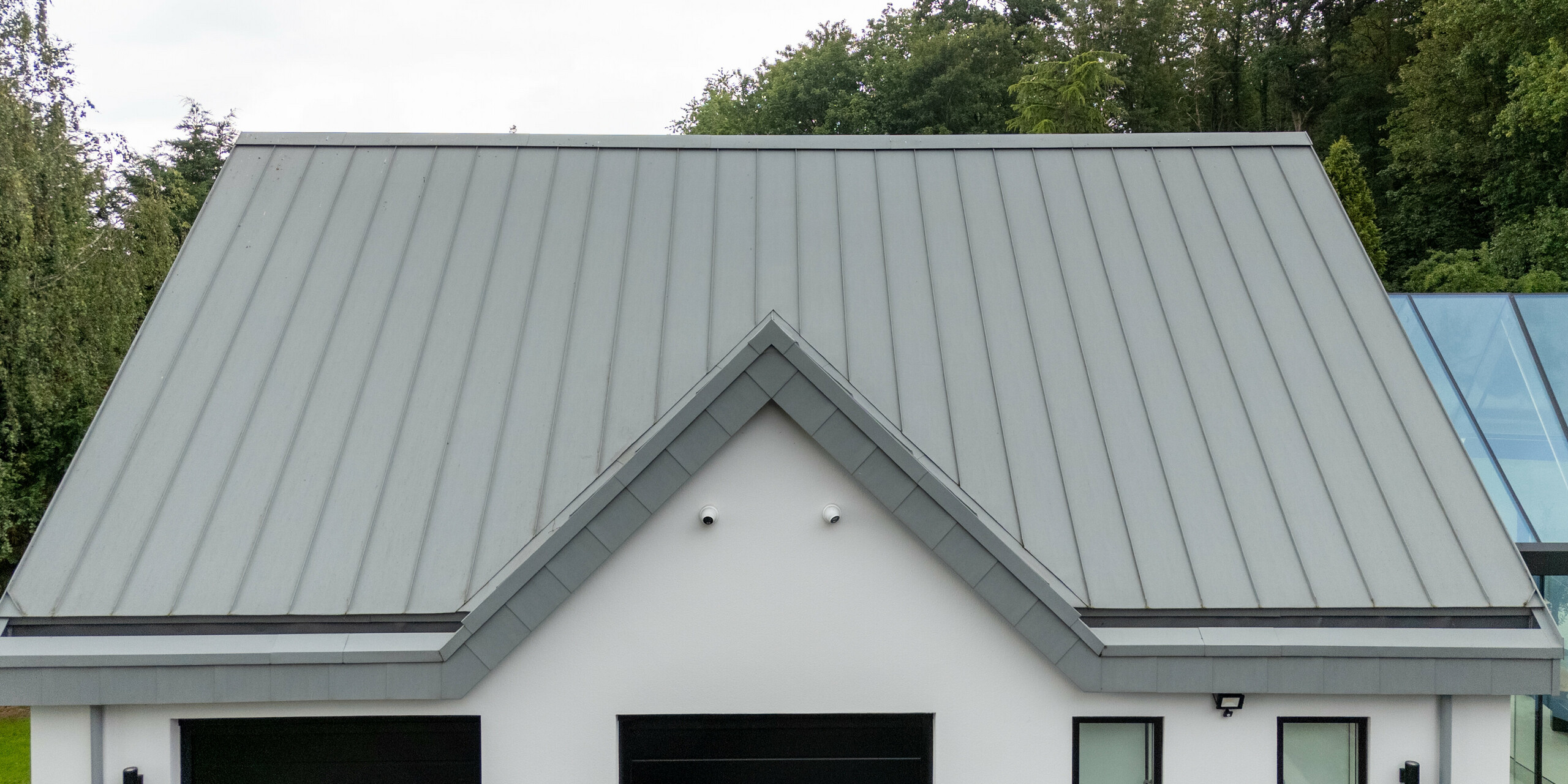 Dieses Bild zeigt eine detaillierte Ansicht des spitzen PREFALZ Aluminiumdachs in Patinagrau mit klaren Stehfalzstrukturen, die das moderne Design der Villa unterstreichen. Die symmetrische Form und die nahtlose Metalloberfläche bieten nicht nur ästhetischen Wert, sondern auch eine hohe Langlebigkeit und Witterungsbeständigkeit. Die perfekte Kombination aus Funktionalität und Stil wird durch die harmonische Integration des Dachs in die Gesamtarchitektur des Gebäudes verstärkt, das sich ideal in die grüne Landschaft von Draycott In The Clay einfügt.