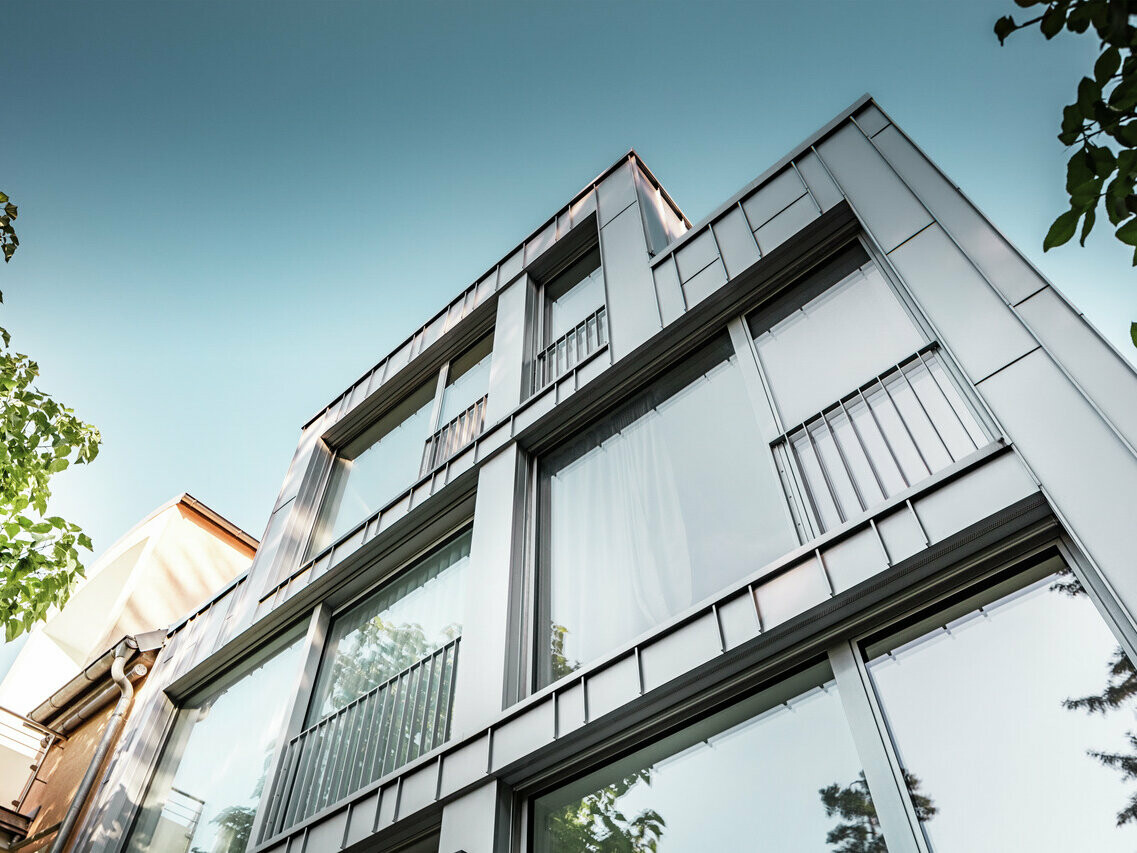 Froschperspektive eines modernen Mehrfamilienhauses mit klarer geometrischer Architektur, gekennzeichnet durch eine glänzende silbermetallische Prefalz Aluminiumfassade. Große Fensterfronten reflektieren den blauen Himmel und die umgebende Vegetation. Das Gebäude zeichnet sich durch schmale Balkone mit metallenen Geländern aus, die ein minimalistisches Design unterstreichen.