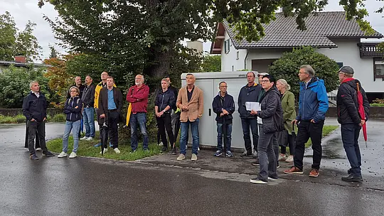 Teilnehmende der PREFA Objekttour versammeln sich an einem regnerischen Tag unter einer imposanten, weit ausladenden Eiche und lauschen den Erläuterungen zu einem sanierten Wohnobjekt.